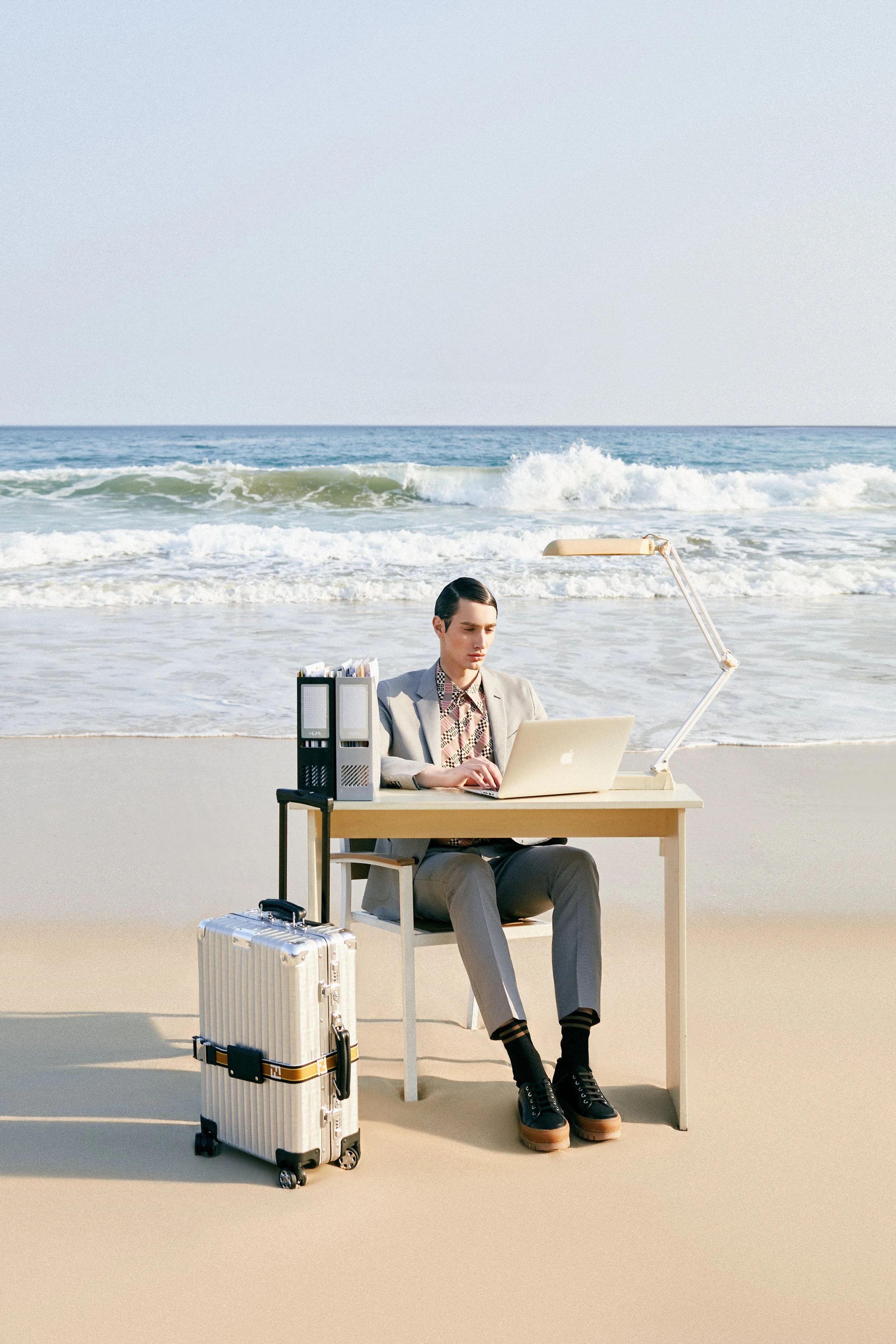 Man working on a laptop at a beach desk with a suitcase beside him, ocean waves in the background.