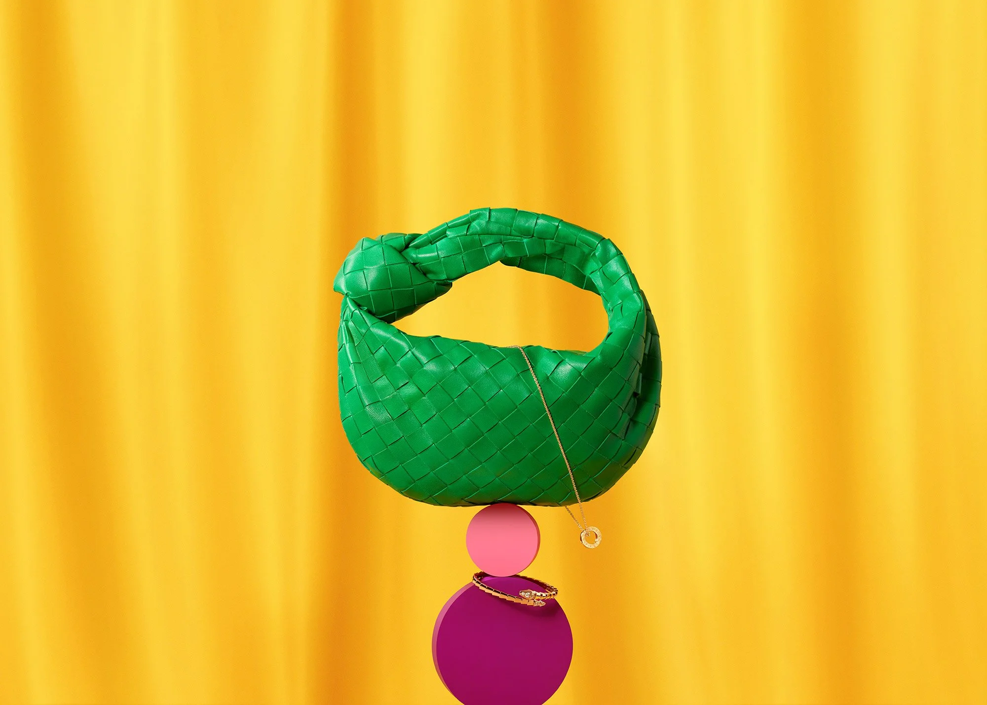 A green woven handbag shaped like a knot, placed on a pink sphere with a purple sphere beneath it, against a yellow curtain background. A gold necklace is draped over the pink sphere.
