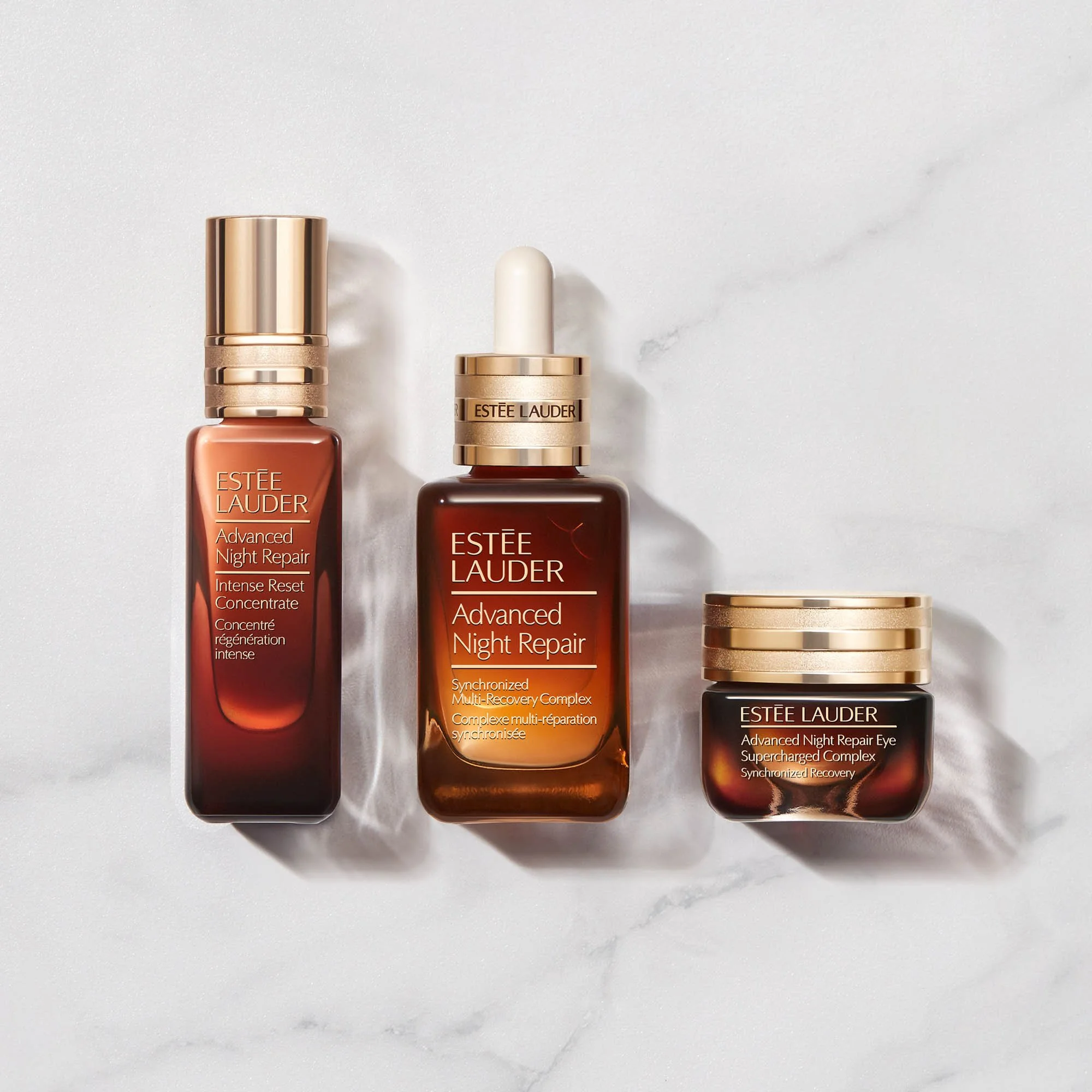 Three Estee Lauder skincare products from the Advanced Night Repair line on a white surface. The first is a tall, amber-colored bottle with a rose gold cap, the second is a smaller jar with a gold cap, and the third is a short jar with a gold cap.