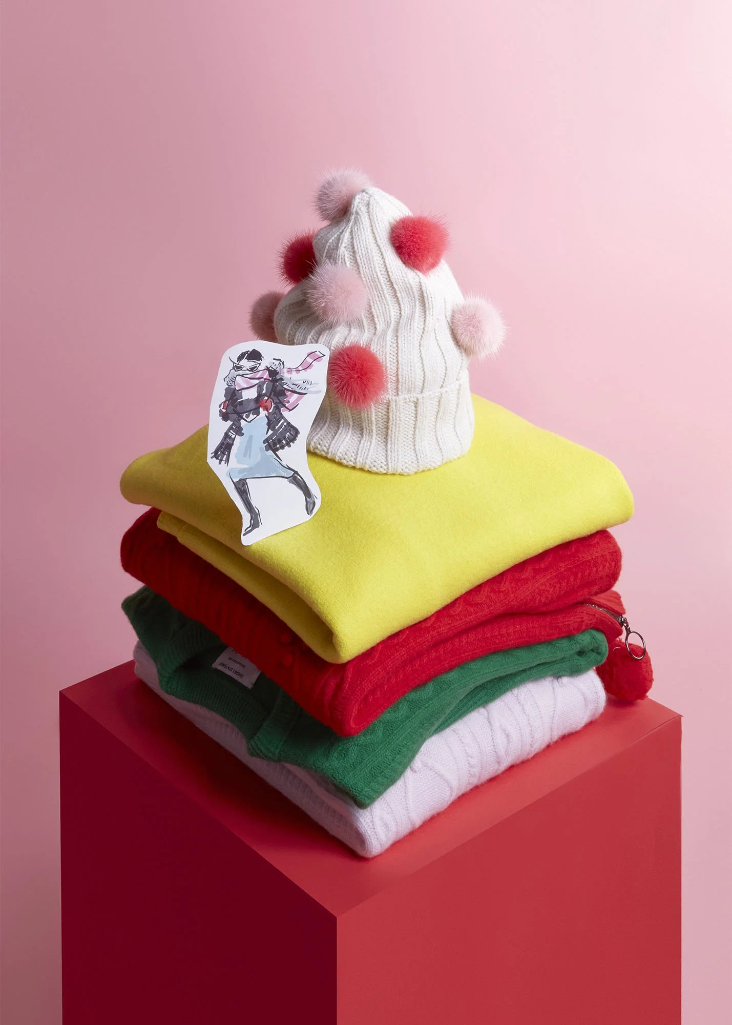 Stack of folded colorful sweaters and a white knit hat with red and pink pom-poms, placed on a pink podium against a pink background.