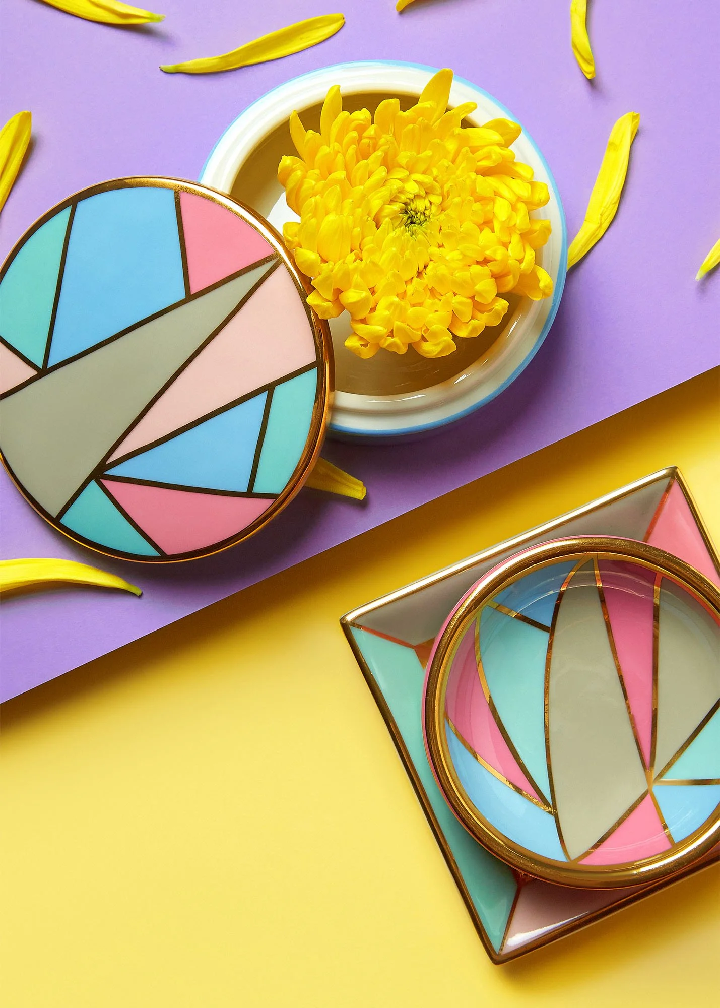 Decorative colorful jewelry boxes with pastel geometric patterns, a yellow flower in a white bowl, and scattered yellow flower petals against a purple and yellow background.