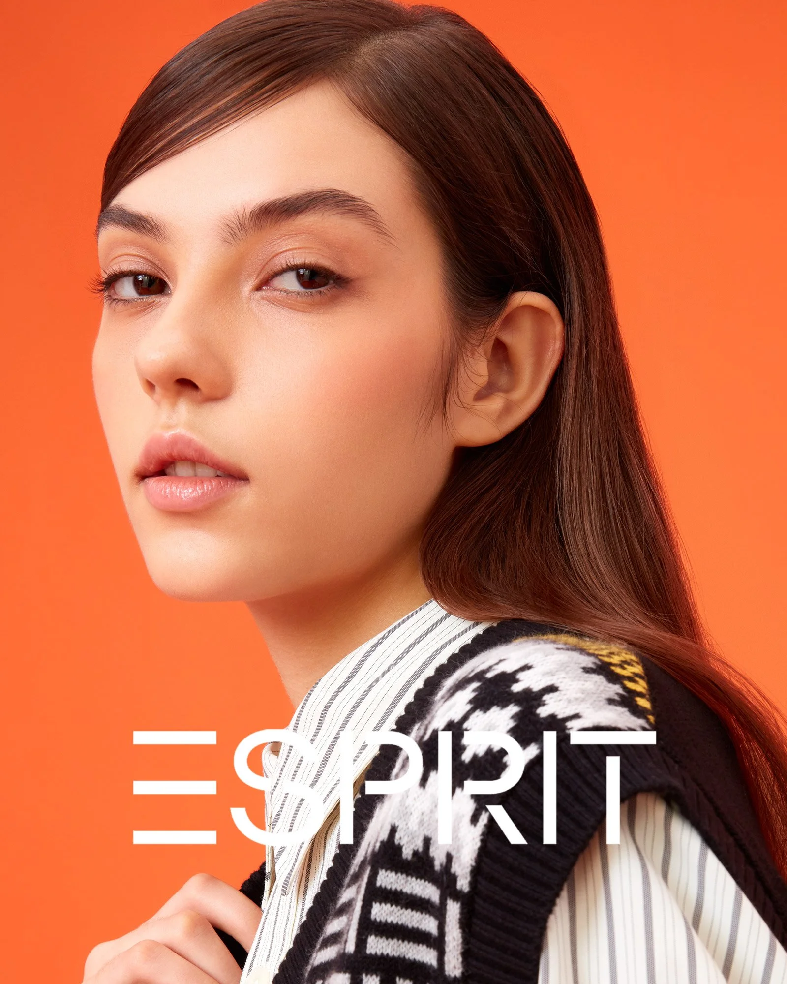 Close-up of a young woman with long brown hair, wearing a striped shirt and patterned cardigan, against an orange background. The word 'ESPRIT' appears in white text at the bottom of the image.