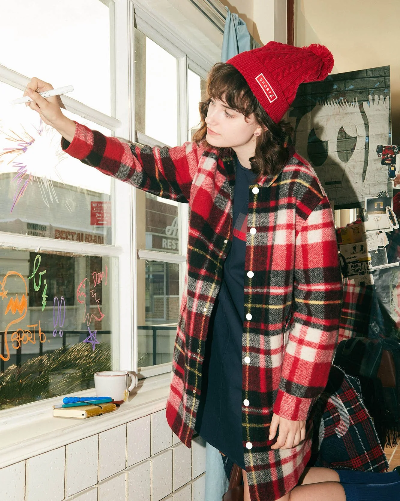 A young woman in a red and black plaid coat and red knit beanie decorating a window with colorful Halloween drawings.
