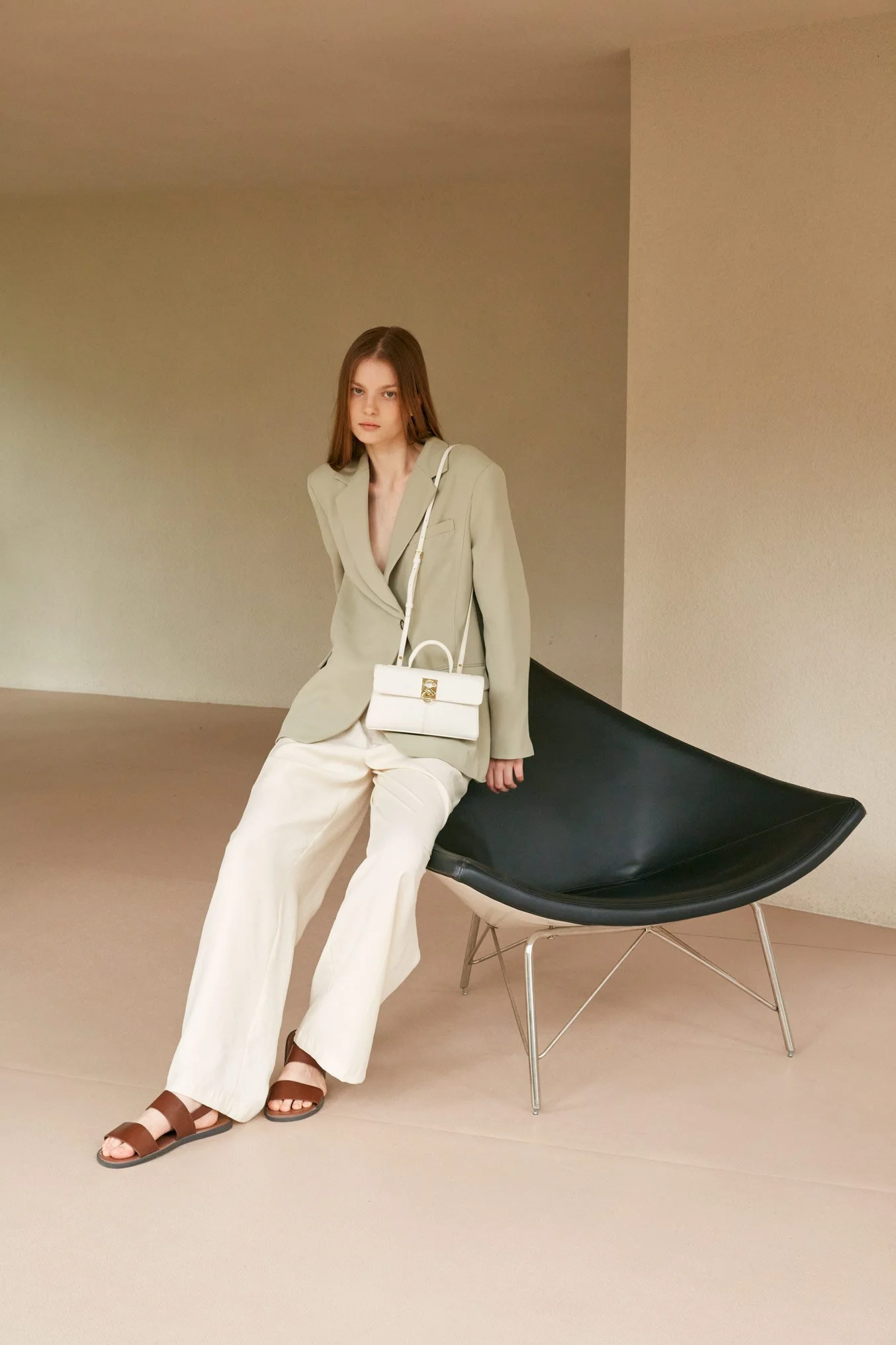 A woman with long brown hair sitting on a black modern chair in a neutral-colored room, wearing a beige blazer, white wide-leg pants, brown sandals, and carrying a white handbag.