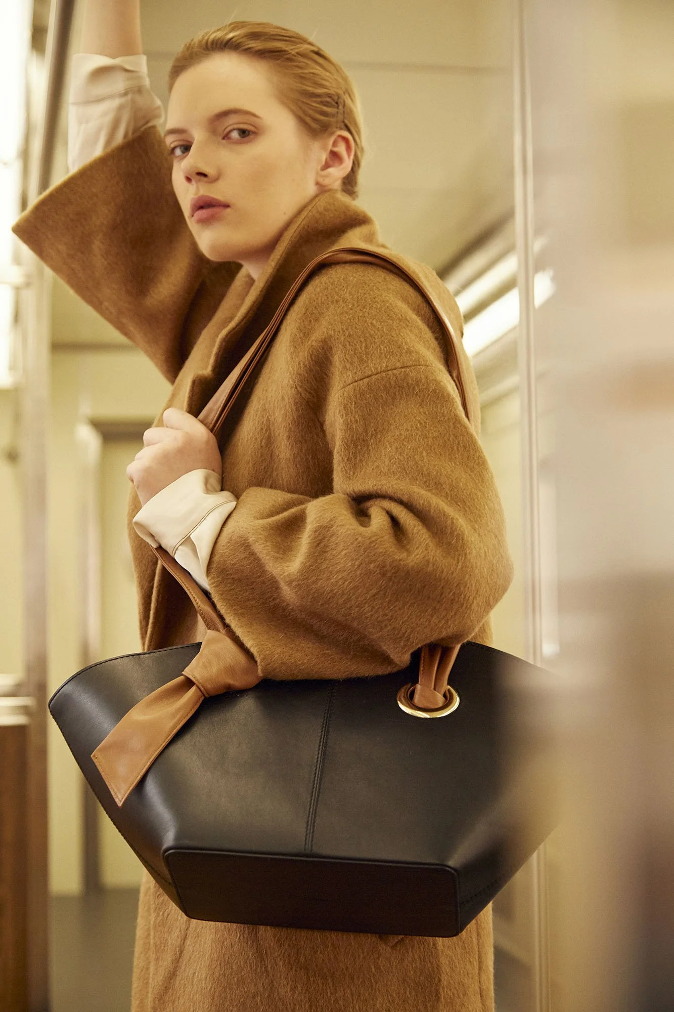 A woman with short red hair and fair skin looking over her shoulder in a brown coat, holding a brown purse and standing in an indoor setting that resembles a train or subway car.