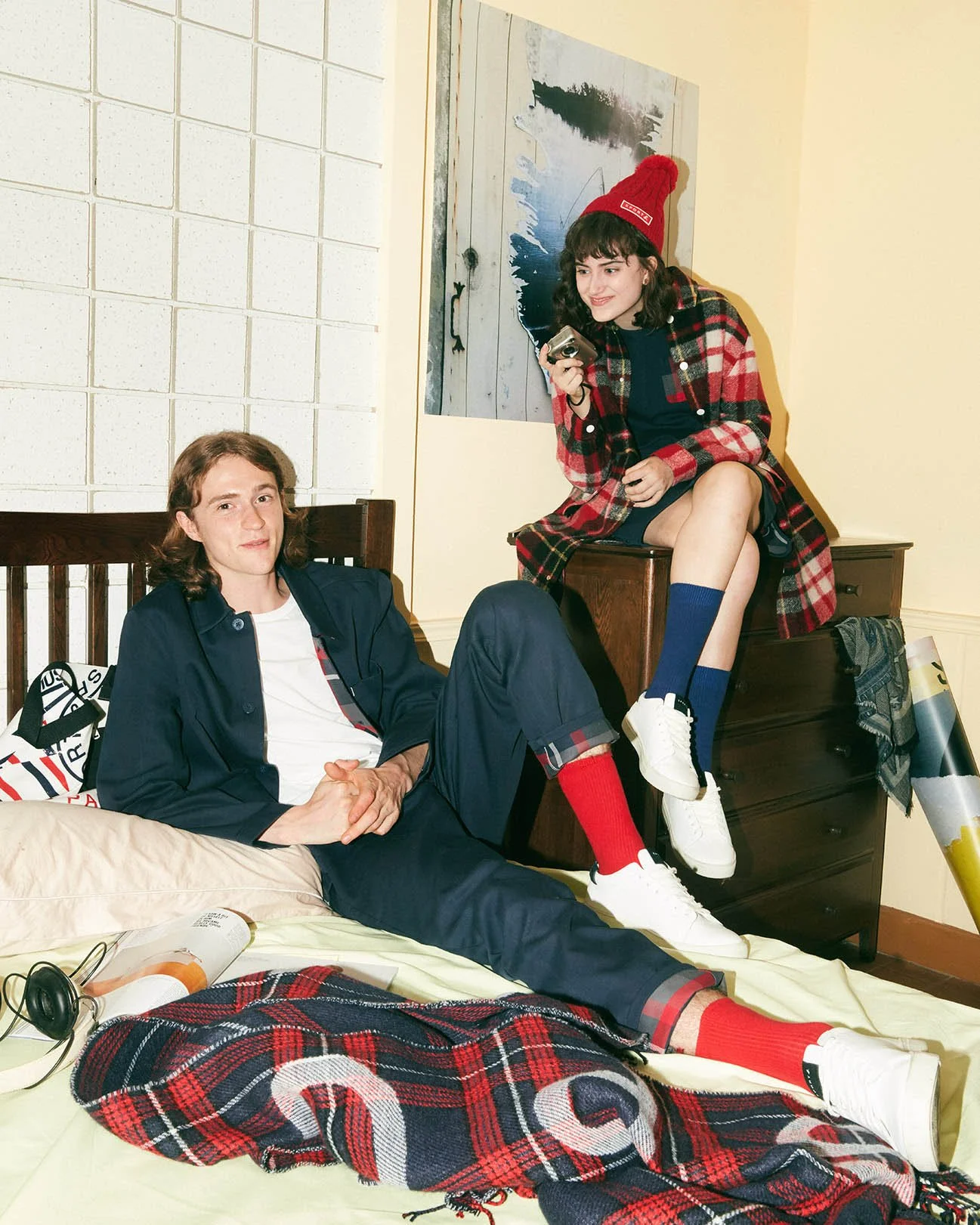 A young man sitting on a bed with a young woman sitting on a dresser next to him. Both are dressed casually with matching plaid patterns, and the woman is wearing a red beanie. They appear to be in a bedroom with a painting on the wall and various it