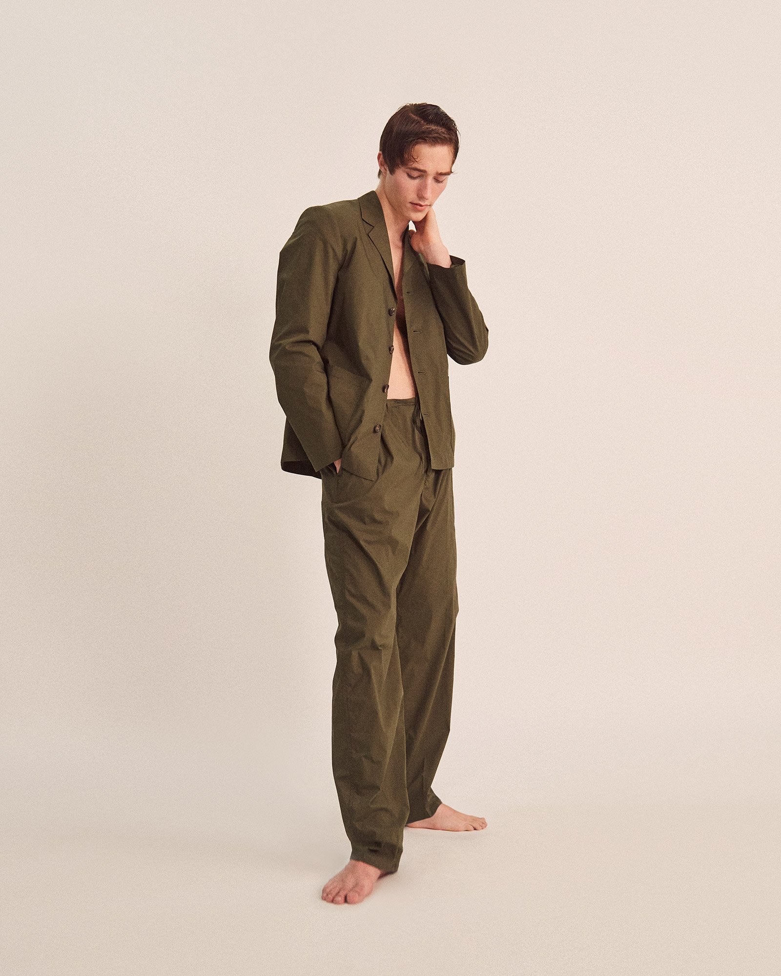 A young man in a green suit with no shirt, standing barefoot against a plain beige background, looking down with one hand on his neck.