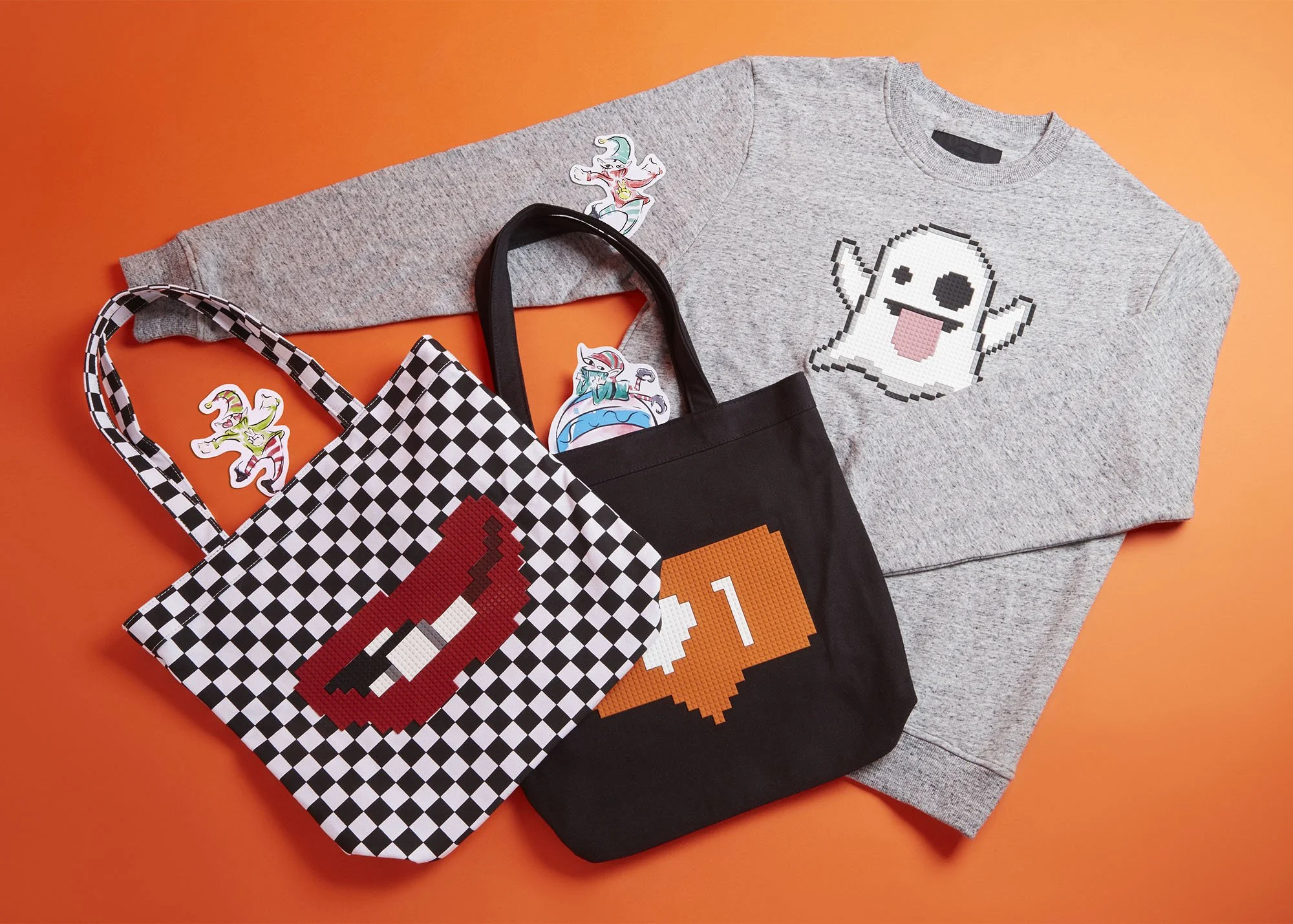 Gray sweatshirt with a pixelated ghost graphic, black and white checkered tote with a red pixelated mouth graphic, orange and white pixelated heart graphic on a black tote, and colorful stickers of a unicorn and a dragon on an orange background.