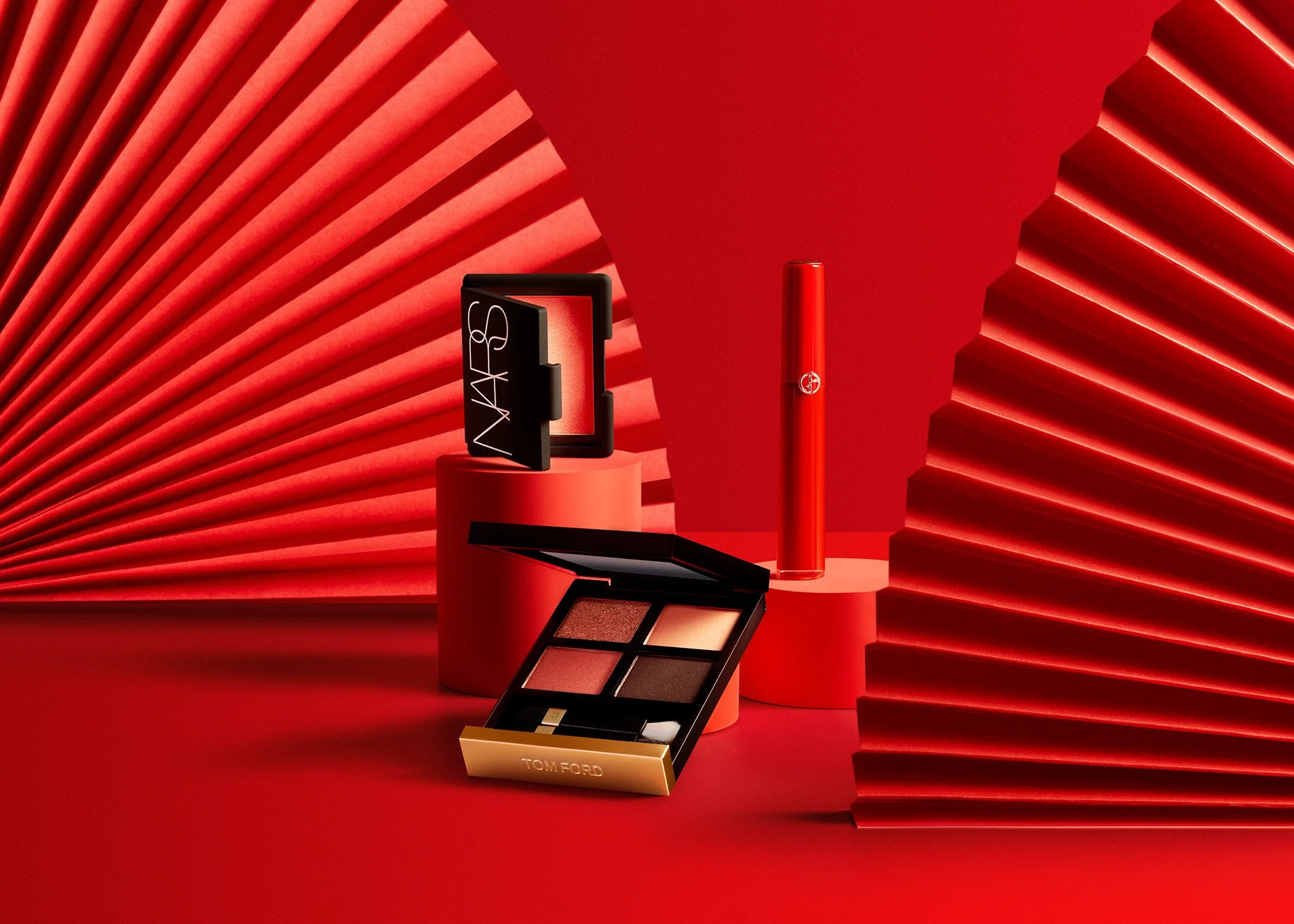 Makeup products including eyeshadow palette, lipstick, blush compact, and a cosmetic case displayed on red cylindrical platforms with red background and textured red paper accents.