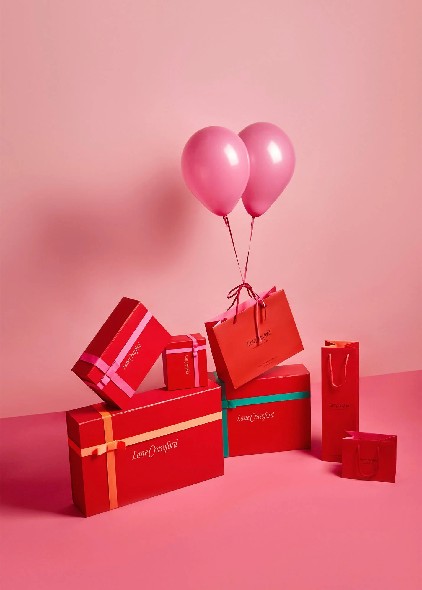 Pink background with two pink balloons tied to gift bags and boxes. The gifts are branded with 'Lane Crawford' and are decorated with colorful ribbons.