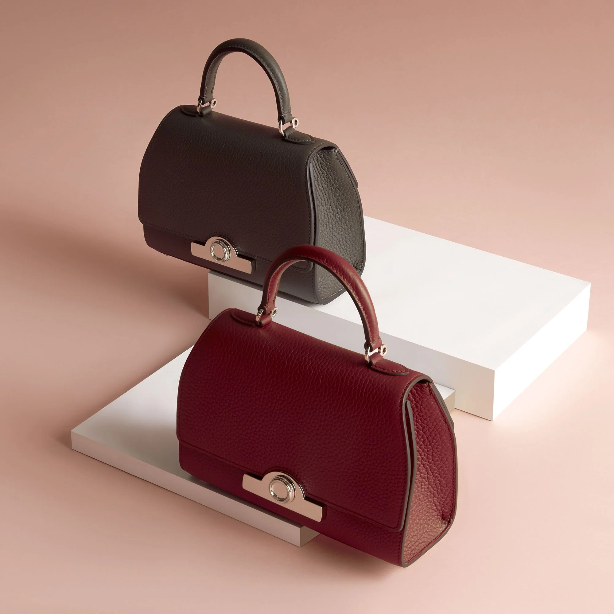 Two designer handbags, one black and one red, placed on white display blocks against a pink background.