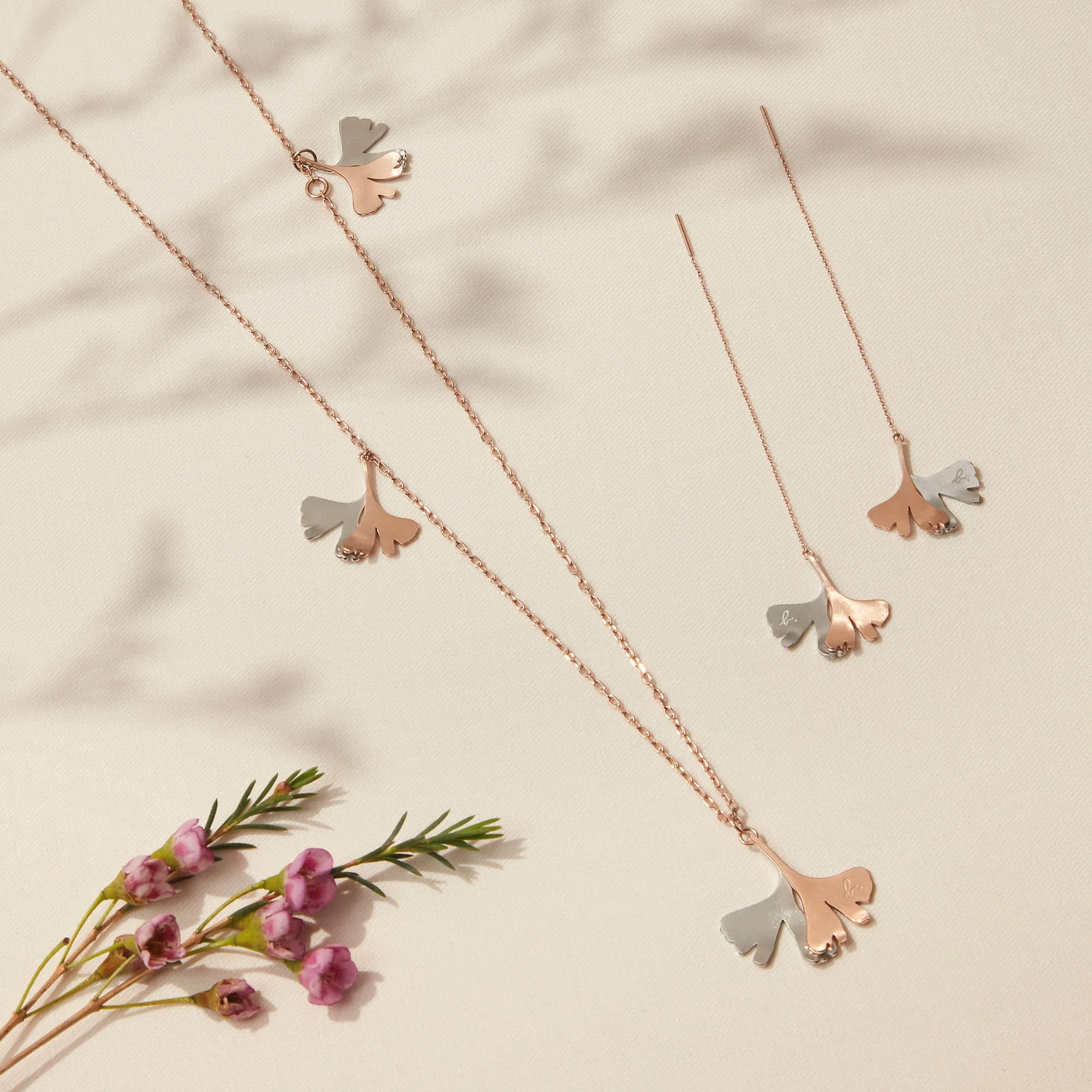 Jewelry set including a necklace and earrings with ginkgo leaf designs, placed on a light-colored fabric with a small branch of pink flowers in the lower left corner.