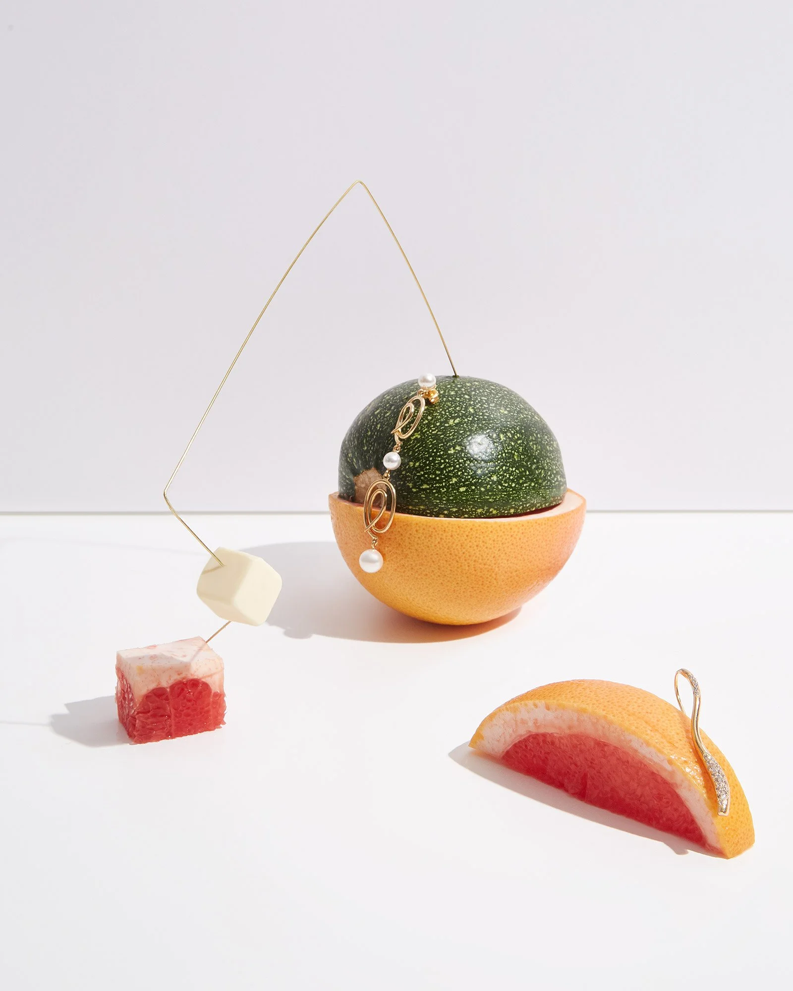 An artistic composition of citrus fruits and jewelry. A halved grapefruit and a half-orange with jewelry dangling from the orange. A lime on top of the grapefruit is decorated with earrings and pearls. A white cube and a small red cube are also part 