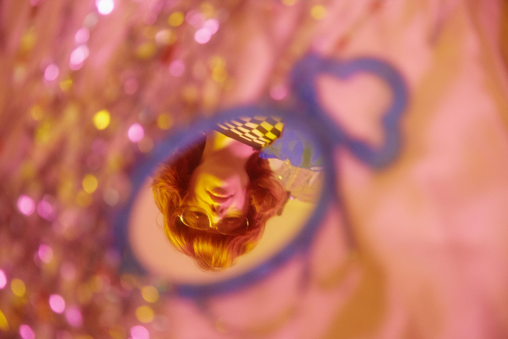 A woman with glasses and red hair looking through a colorful mirror with pink, yellow, and purple lighting, creating a vibrant, bokeh effect.