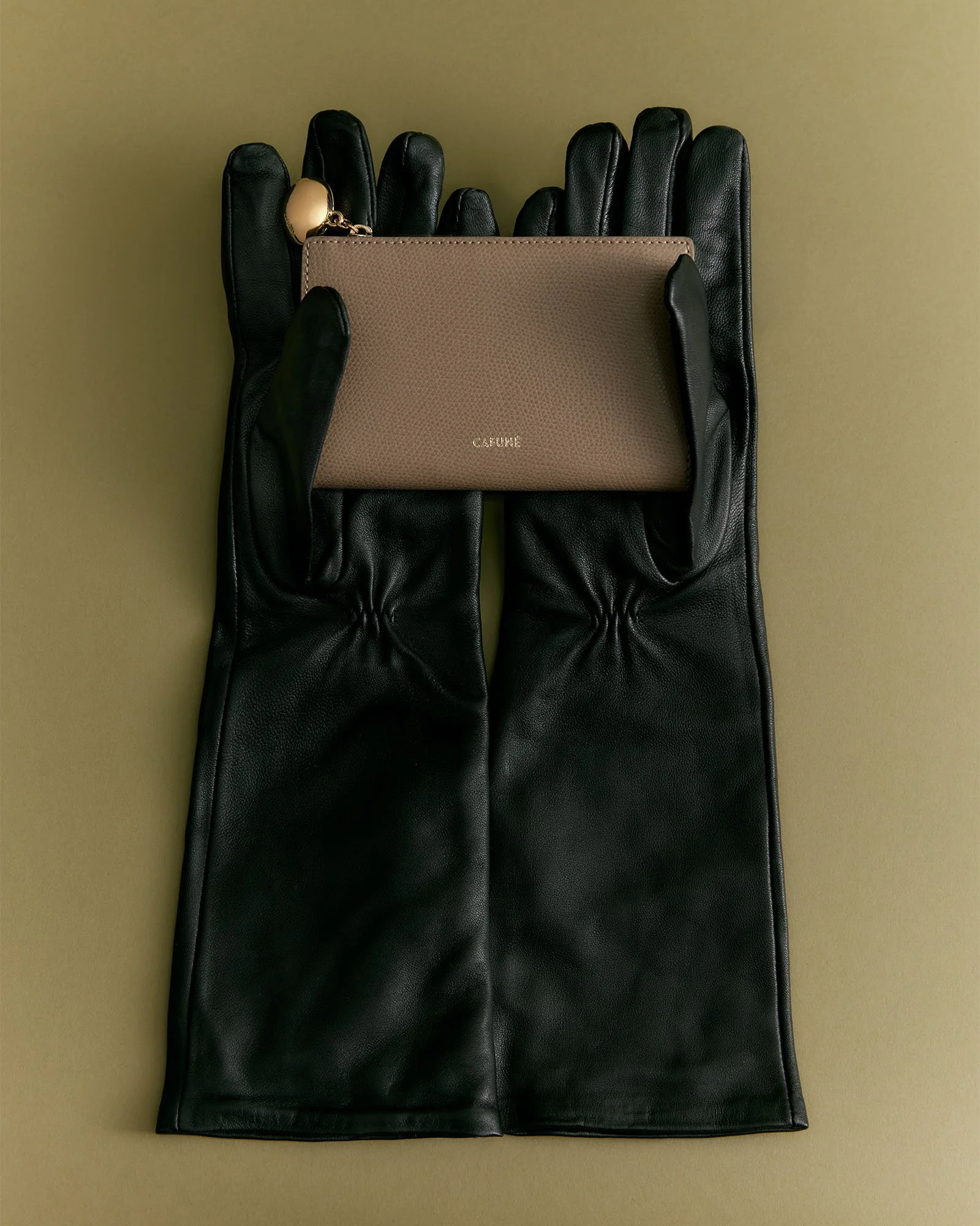 Black leather gloves holding a beige designer wallet with gold hardware on a beige background.