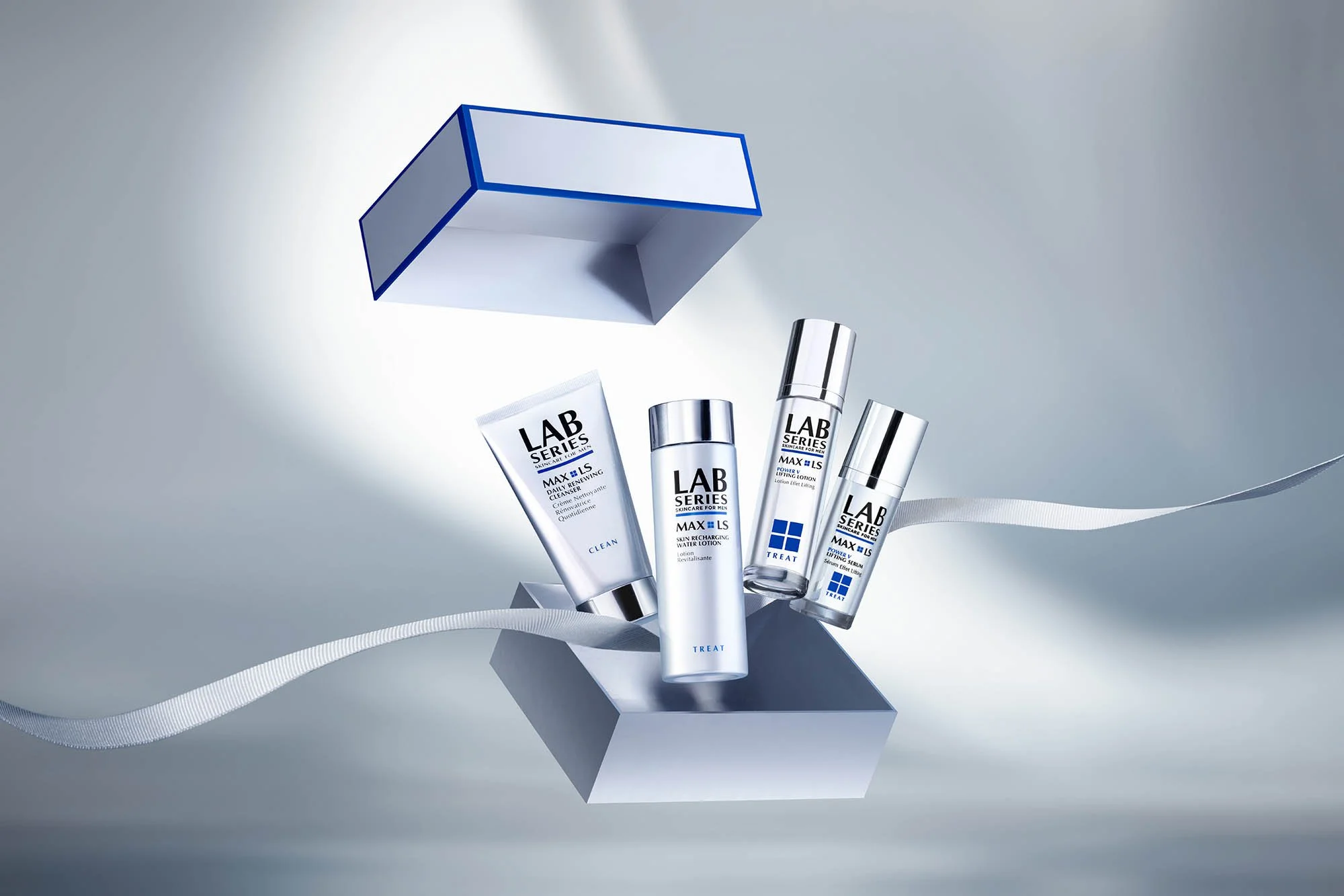 A collection of skincare products from the Lab Series Max LS line, including a cleanser, skin recharging water lotion, and other items, are displayed on a reflective surface with a ribbon, with some products inside a partially open box.