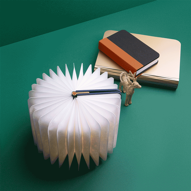Decorative tabletop clock designed to resemble an open book with pages, alongside stacked notebooks and a small figurine of a goat on a green surface.
