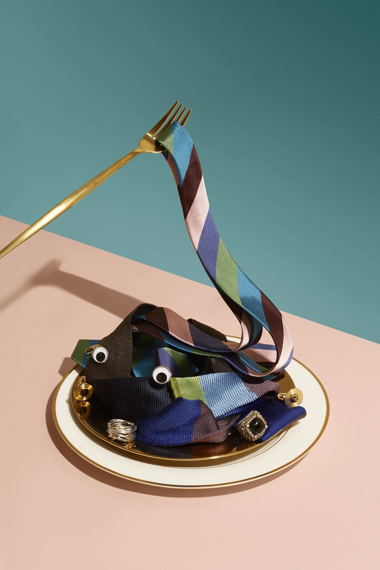 A gold fork with a colorful, patterned ribbon draped over it, resting on a decorative plate with rings and googly eyes, set against a pink and blue background.