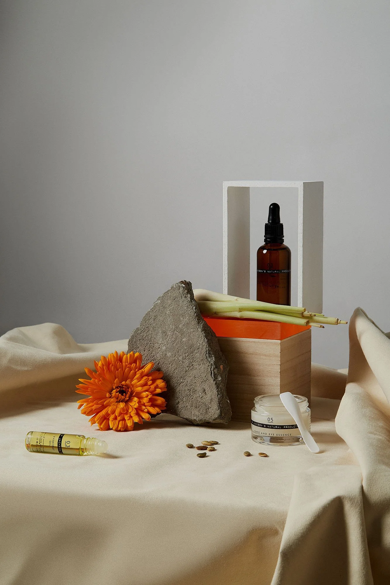Arrangement of skincare products, a flower, wooden blocks, and a rock on a beige draped surface with a plain background.