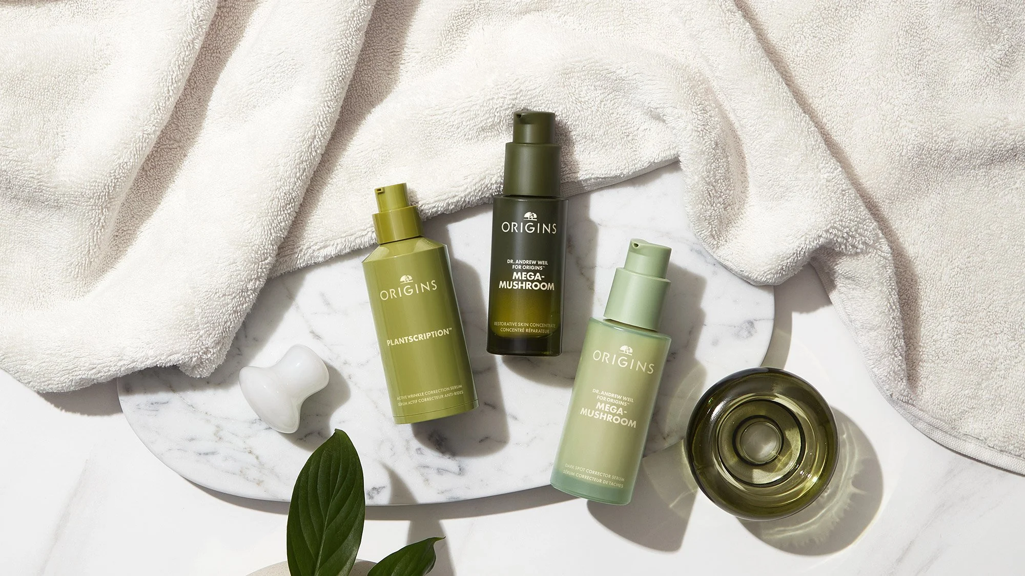 Three bottles of Origins skincare products on a marble tray, with a towel, a jade roller, and a small green glass container nearby.