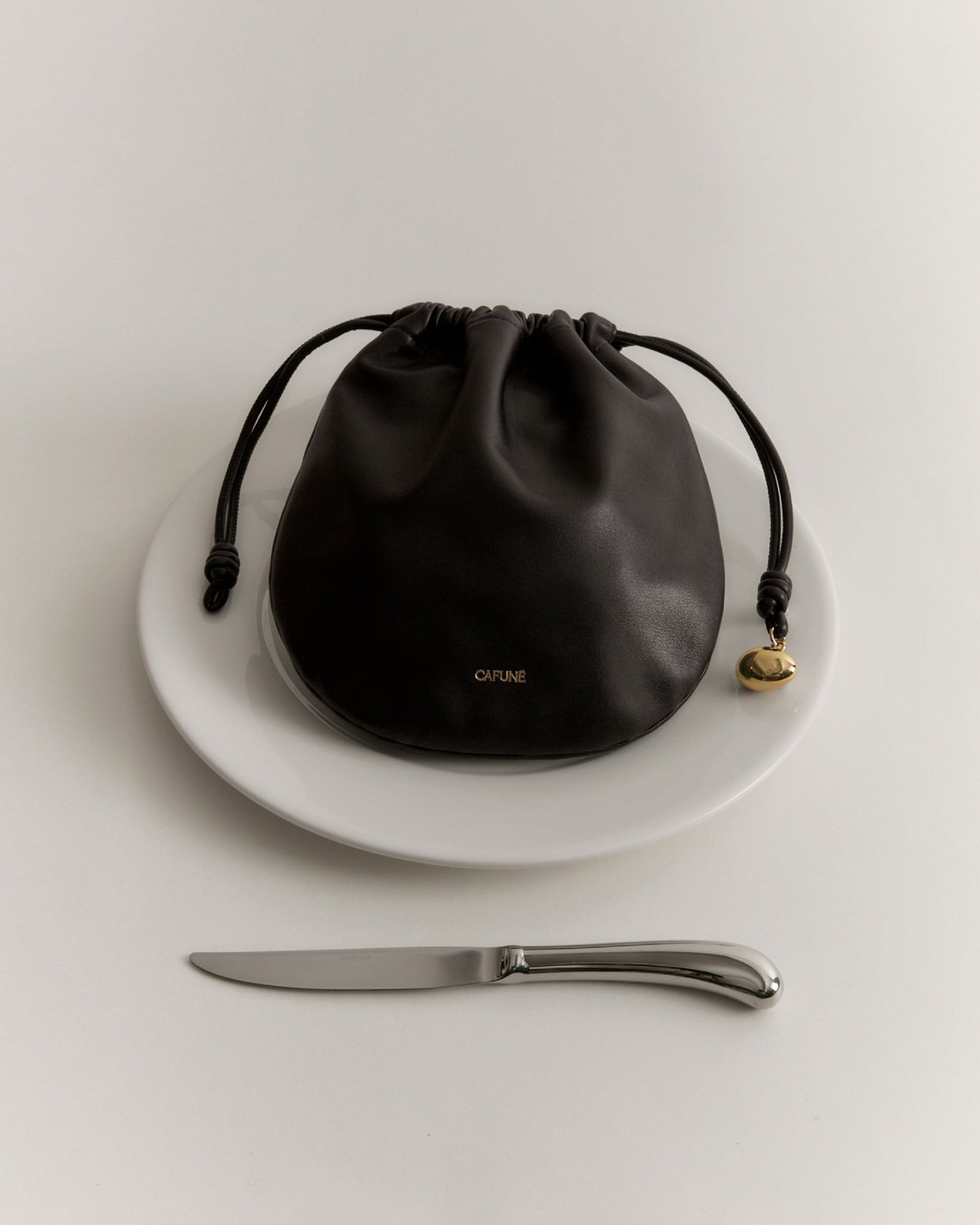 A black drawstring pouch with gold text reading 'CAFUNE' on a white plate, placed on a white table with a silver knife underneath.