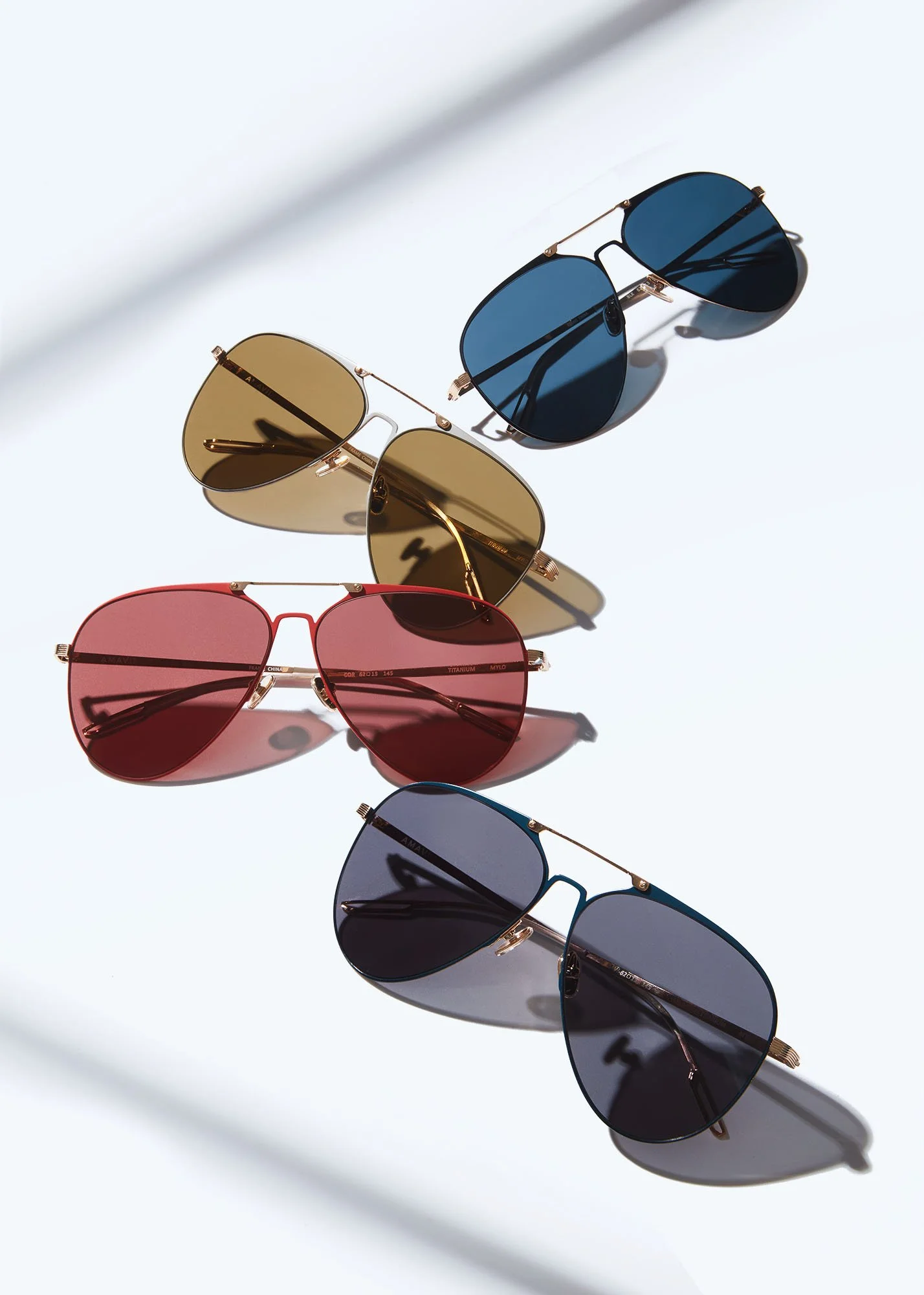 Four pairs of sunglasses with different colored lenses - gold, blue, pink, and dark tinted - arranged on a white surface with shadows.