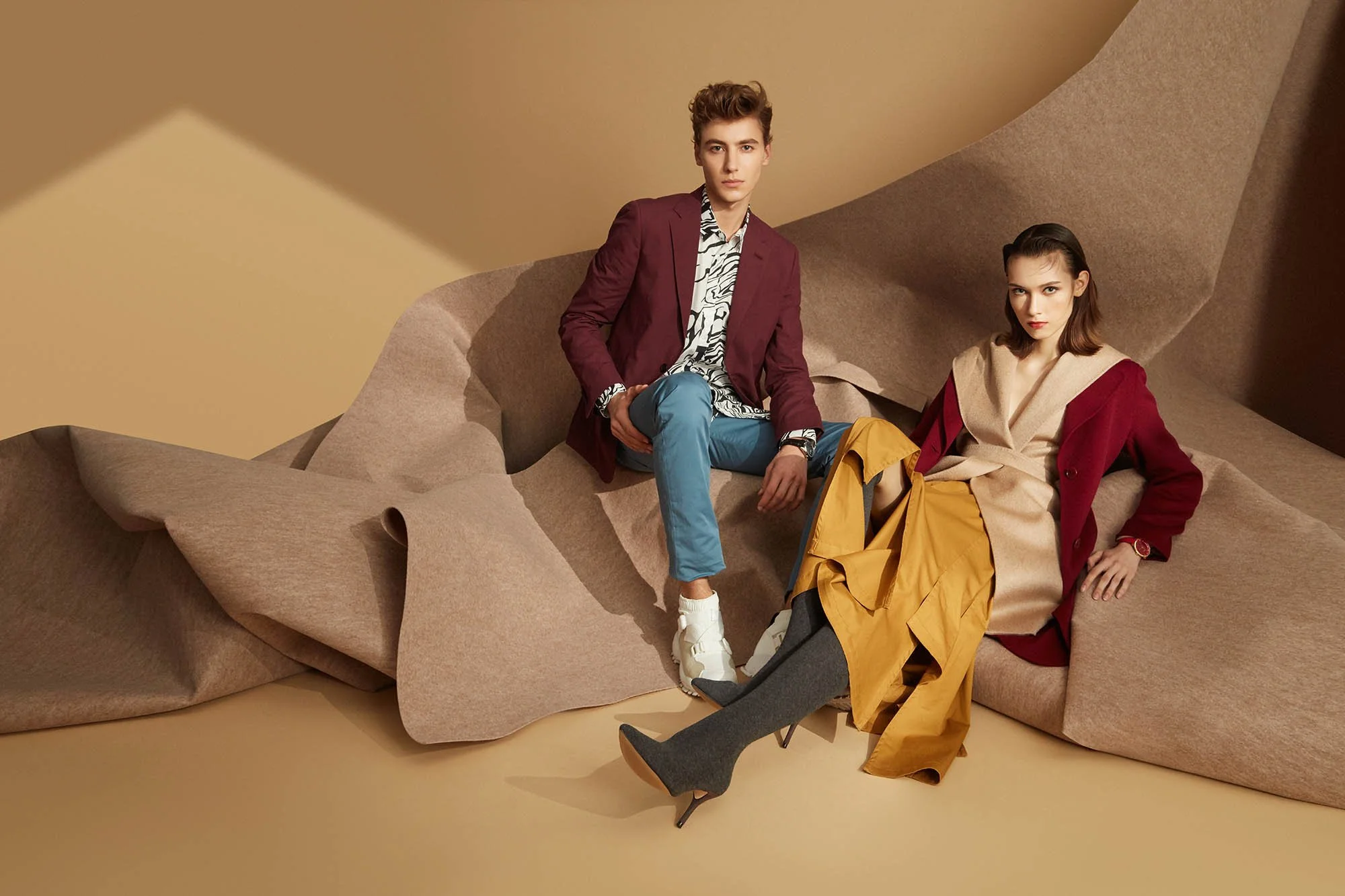Two young adults, a man and a woman, sitting on large crumpled brown paper. The man has light skin, short curly hair, and wears a maroon jacket over a patterned shirt with light blue pants and white sneakers. The woman has light skin, long brown hair