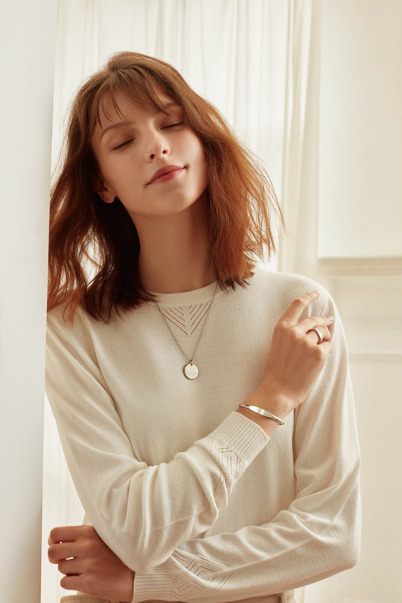 Young woman with shoulder-length red hair smiling gently, wearing a cream sweater, a necklace, bracelet, and ring, standing indoors near a white wall and curtains.