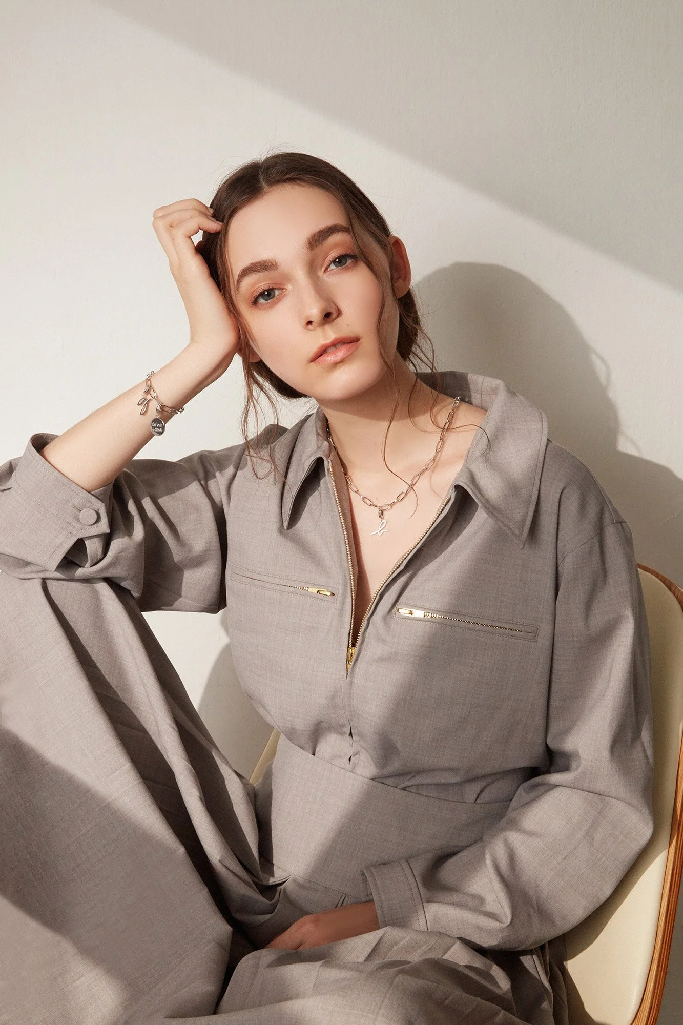 A young woman with light skin, brown hair, and blue eyes wearing a light gray zip-up jacket and matching pants, sitting on a chair against a plain background, looking at the camera with her hand resting on her head.