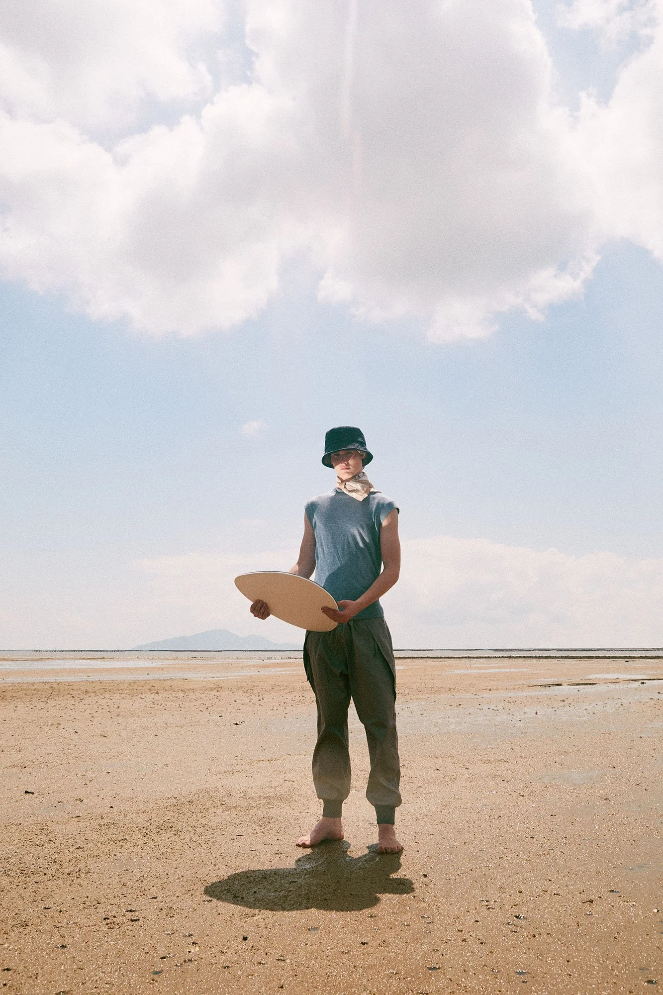 Person standing barefoot on sandy beach, holding an art palette, wearing a black hat, sleeveless shirt, and loose pants, with clouds in the sky and distant mountains.