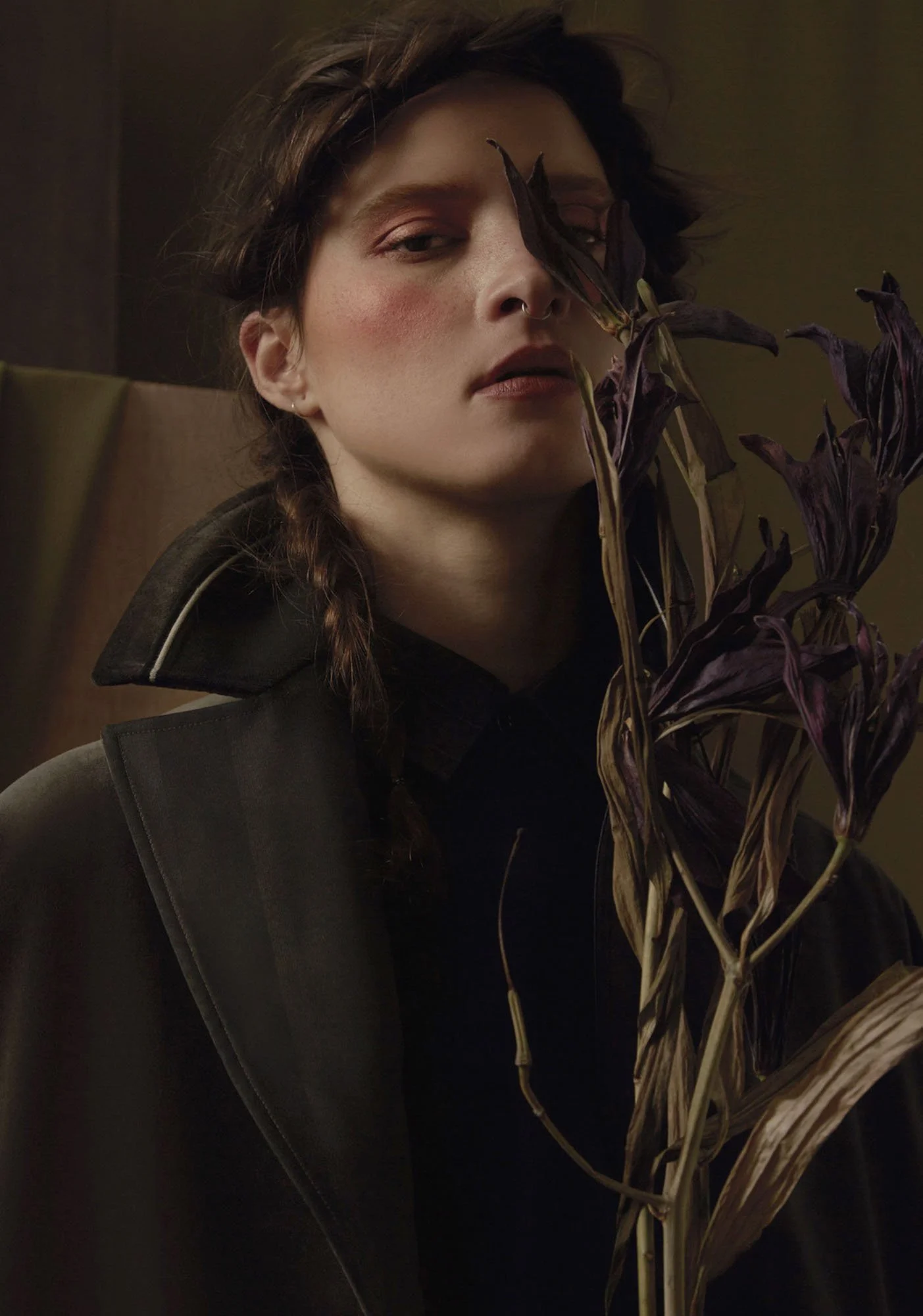 A woman with braided hair and a septum piercing holds dried, dark purple leaves in front of her face, wearing a dark jacket with a high collar.