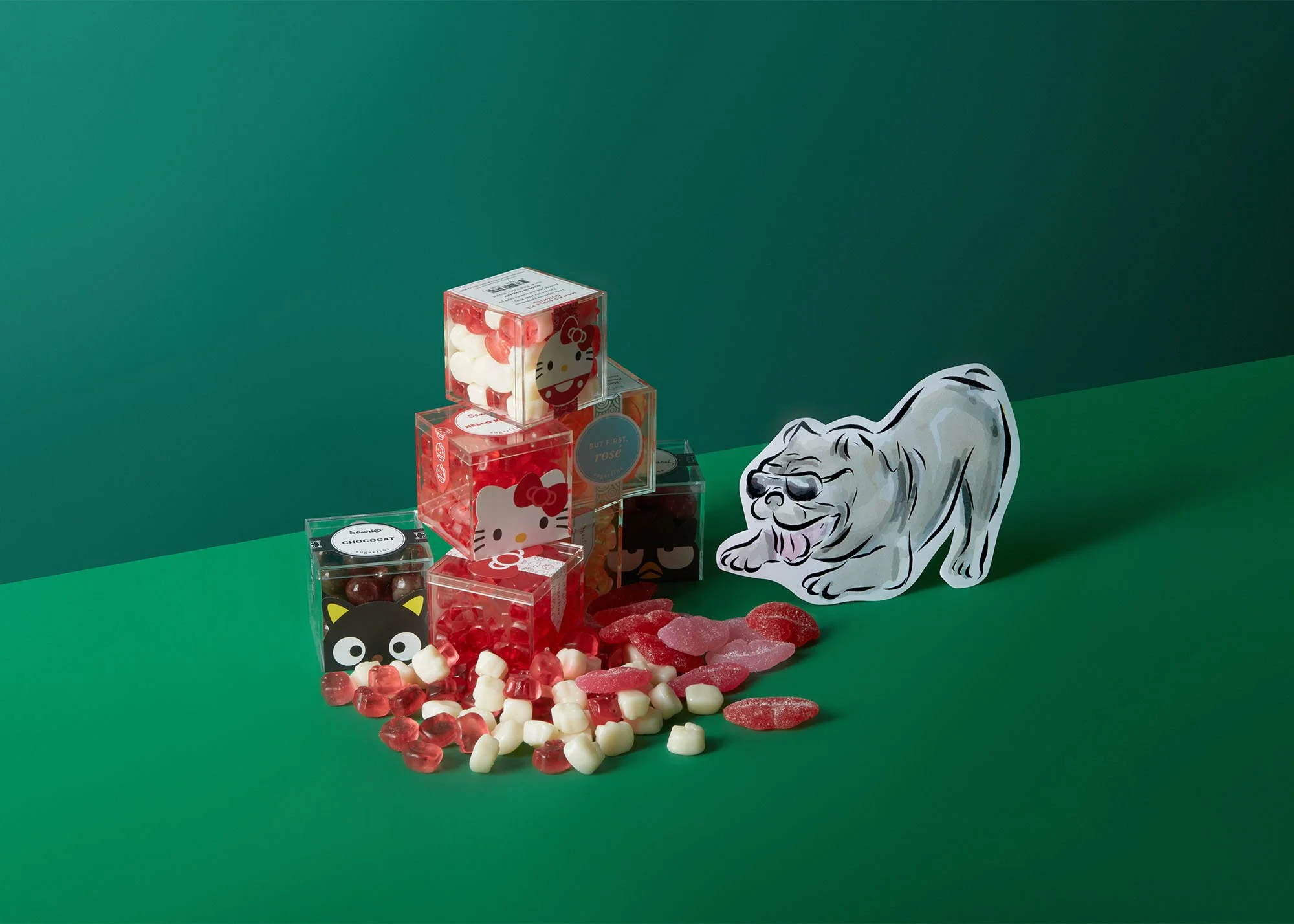 Various Hello Kitty themed candy boxes and gummy candies on a green surface with a paper cutout of a smiling dog.