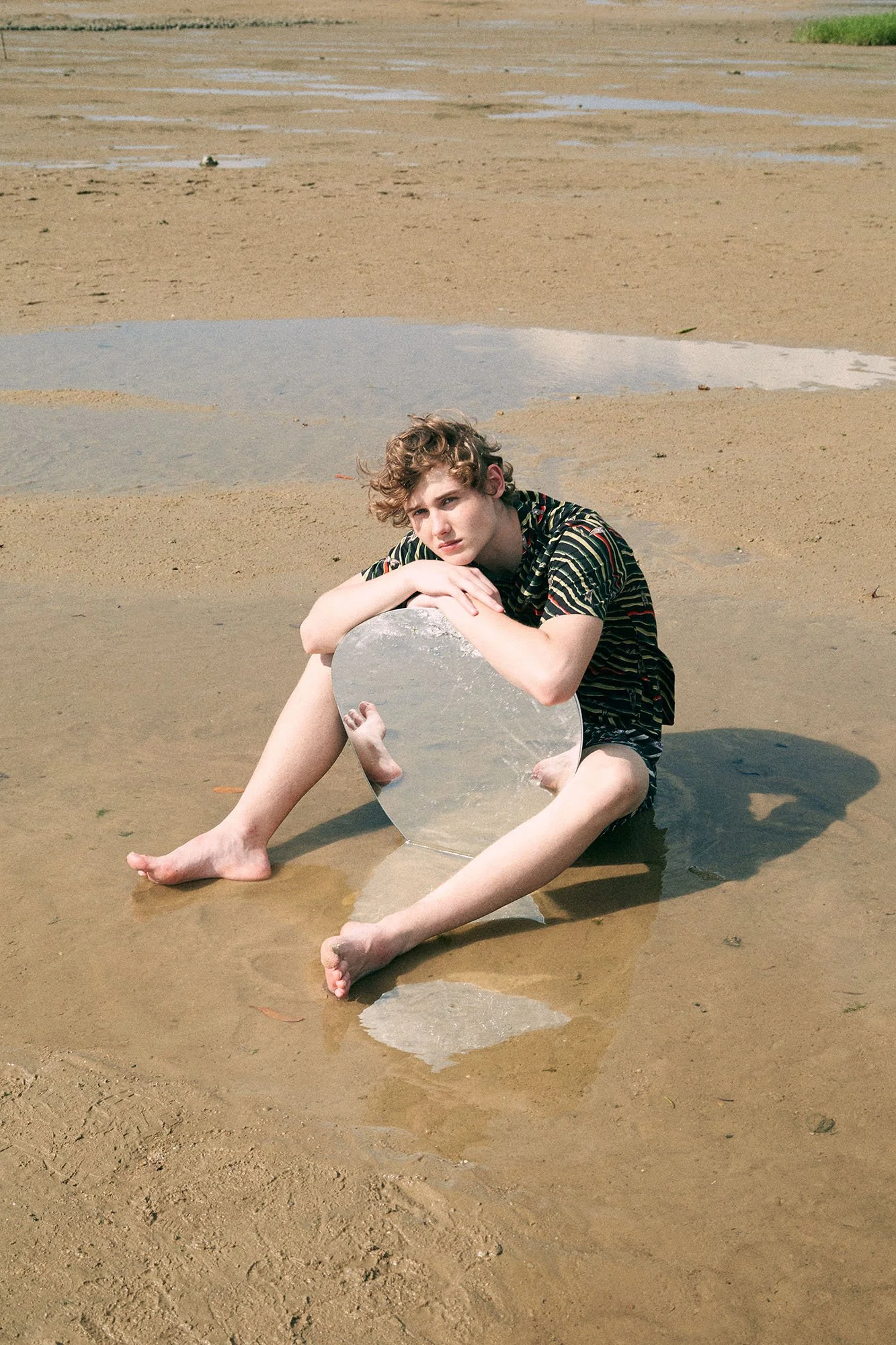 A young man sitting on a sandy beach holding a large broken reflective mirror.