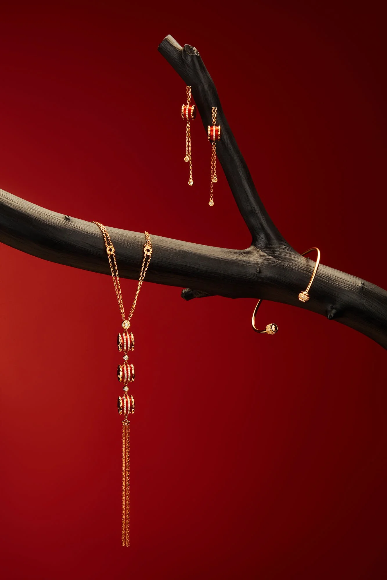 Gold jewelry set including a long necklace, a pair of dangling earrings, a cuff bracelet, and a chain with beads, displayed on a dark branch against a red background.