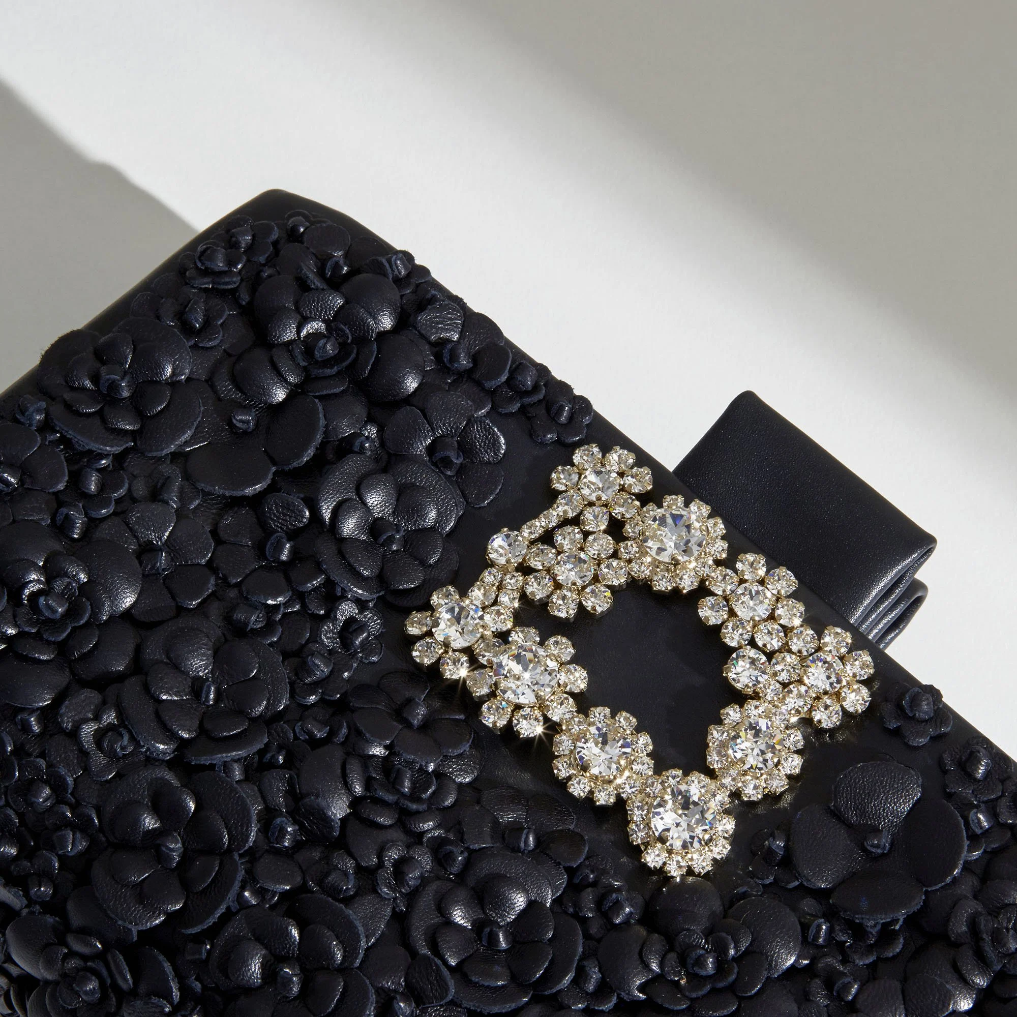 Close-up of a black clutch purse adorned with textured floral embellishments, a sparkling rhinestone buckle, and a black satin ribbon attached.