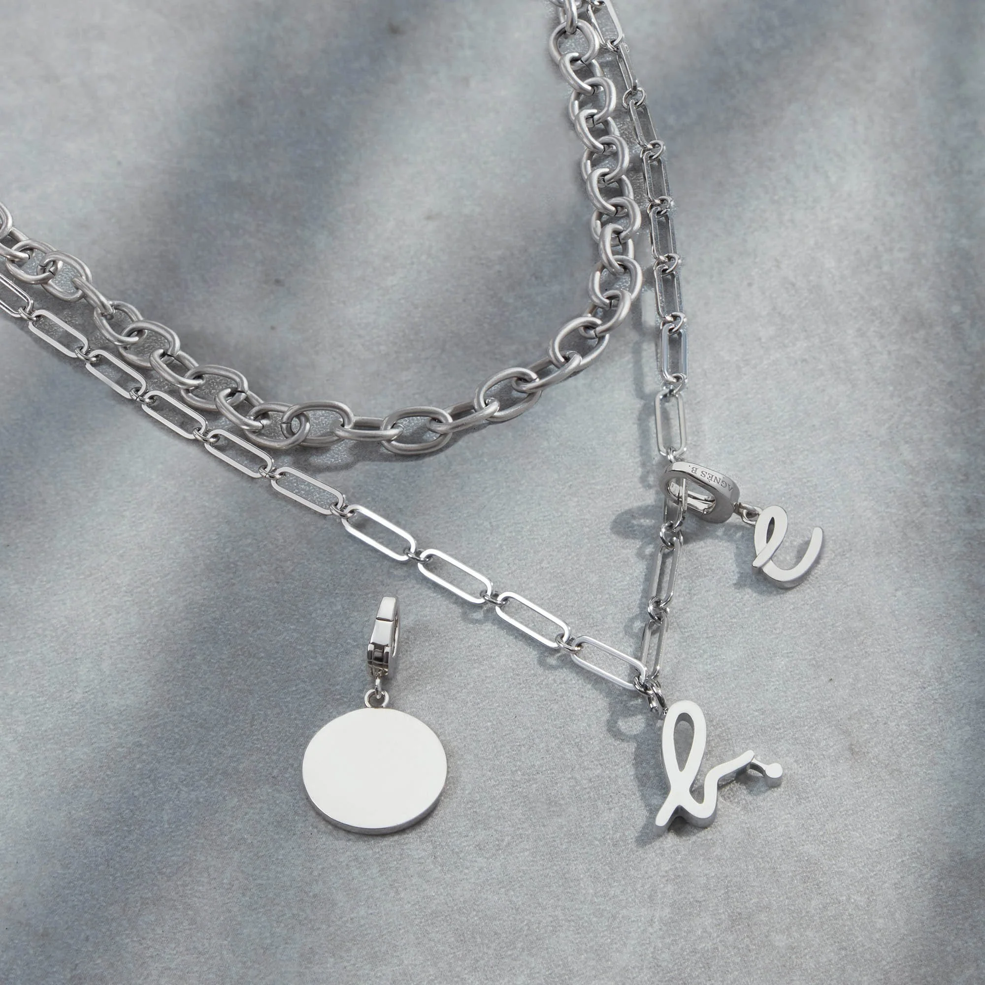 Silver necklaces with letter and pendant charms on a light gray background.