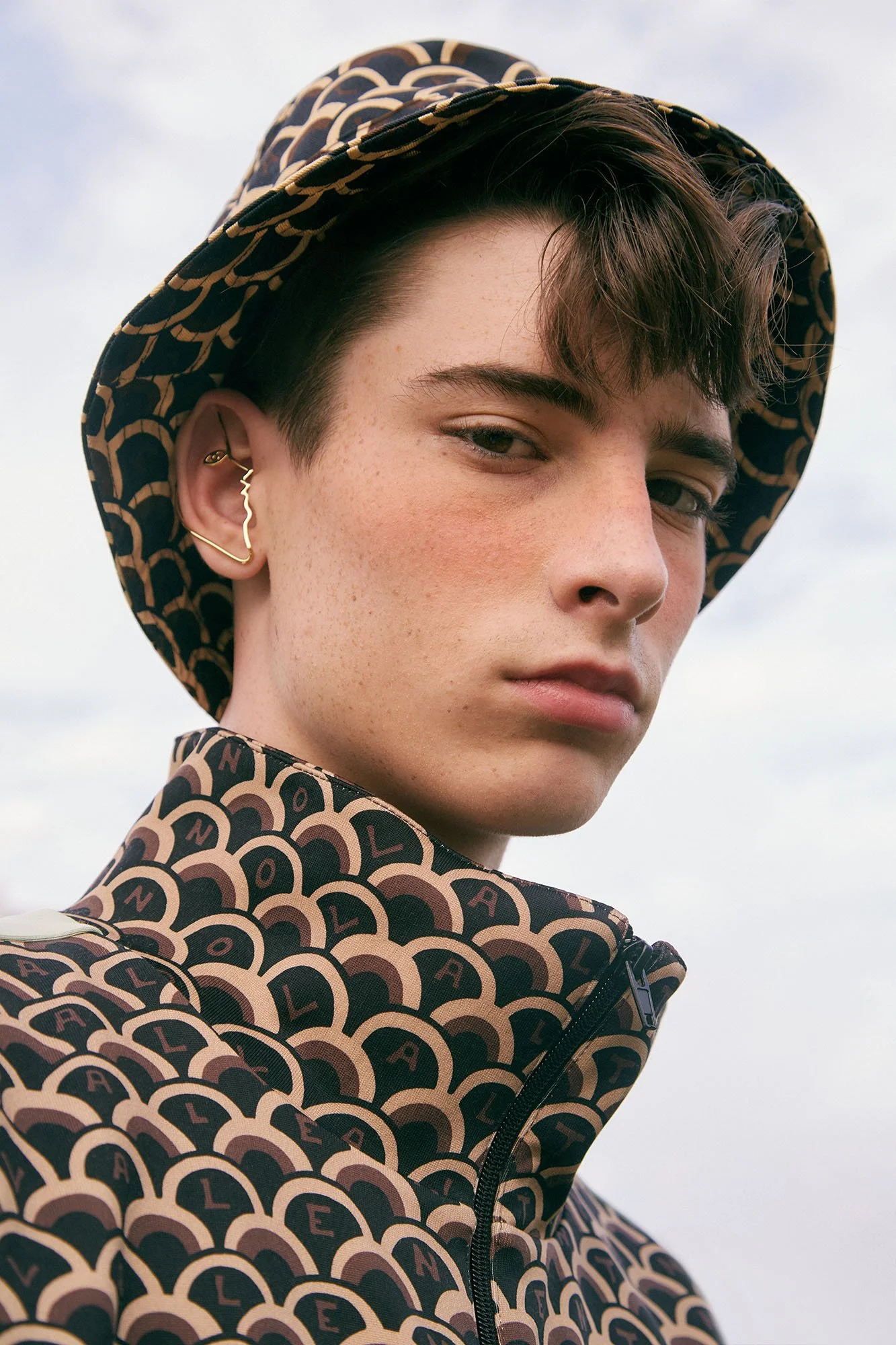 A person wearing a patterned hat and matching jacket with the word 'BALENCIAGA' repeated on it. They have short brown hair, light skin, and gold jewelry in their ear, posing outdoors with a cloudy sky in the background.