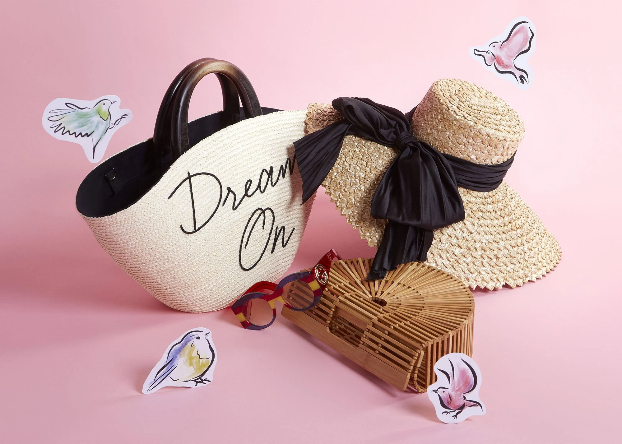 Pink background with a large straw tote bag that has the words 'Dream On' written on it, a wide-brimmed straw hat with a black ribbon, colorful sunglasses, a woven clutch, and drawn bird stickers around.