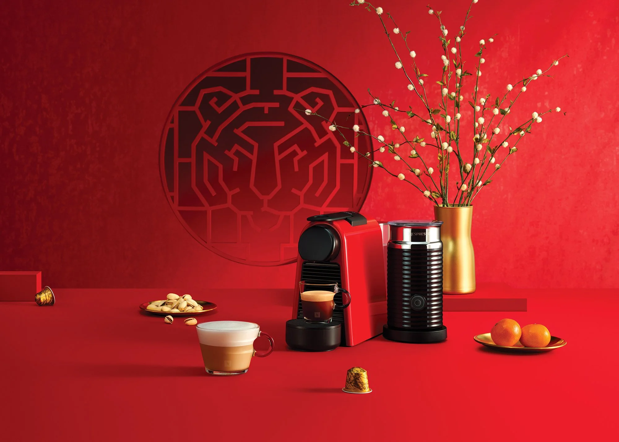 Red coffee machine and a coffee grinder on a red table, with cups of coffee, a bowl of pistachios, oranges, a vase with white flowers, and a decorative wall panel with a lion's face design in the background.