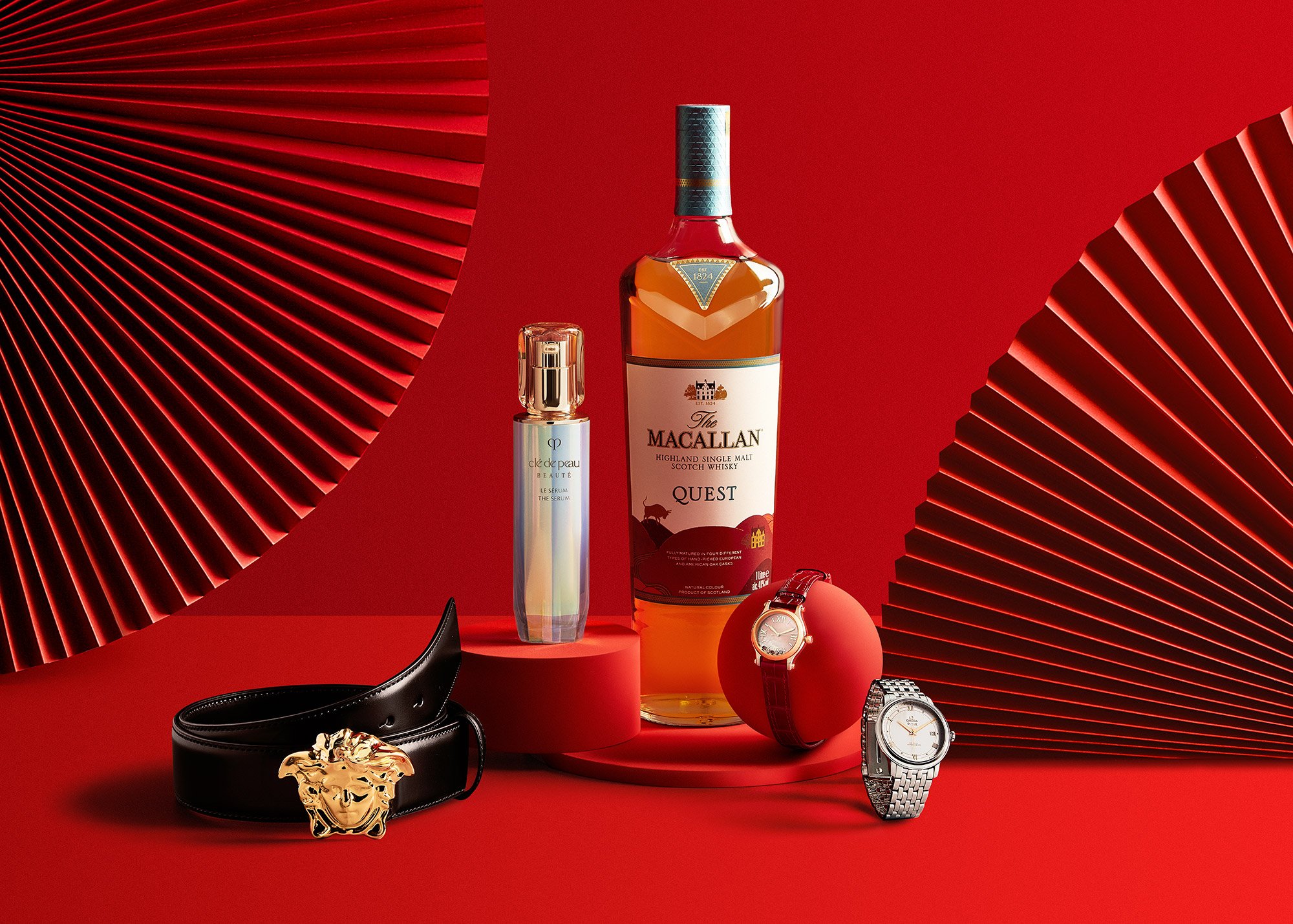 A bottle of The Macallan Quest Highland Single Malt Scotch Whisky, a perfume spray, a wristwatch with a leather strap, a silver bracelet watch, a black belt with a lion's head buckle, and a red ball, all set against a red background with large red pl