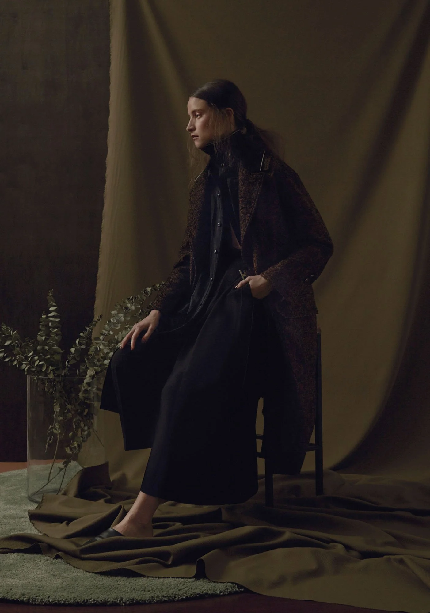 A woman with long brown hair sitting on a black chair against a dark brown backdrop, wearing a dark coat and black pants, with a cardboard of leaves on the surface next to her.