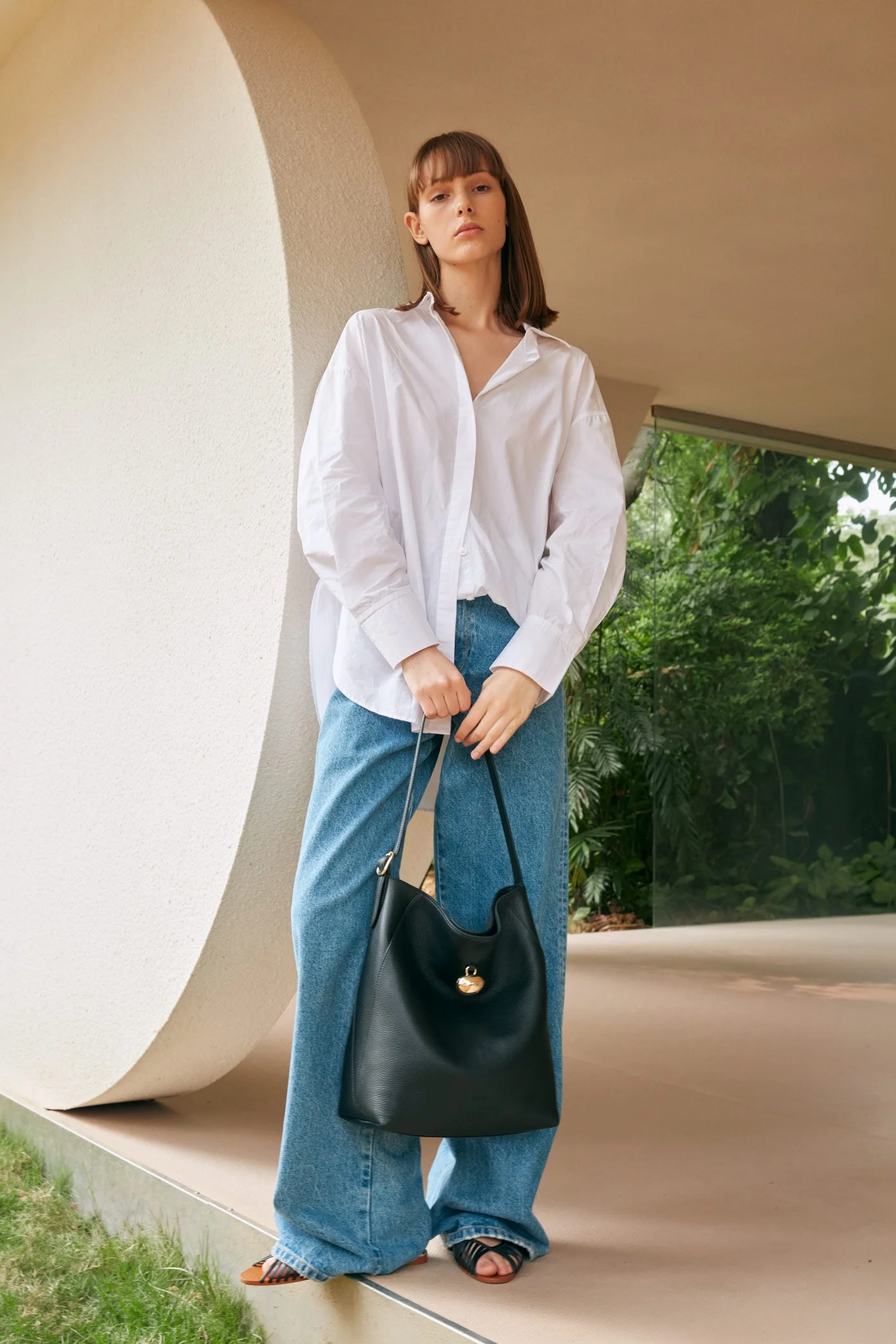 A young woman with shoulder-length brown hair wearing a white button-up shirt and blue jeans standing outdoors next to a curved architectural wall, holding a black handbag, with green foliage visible in the background.