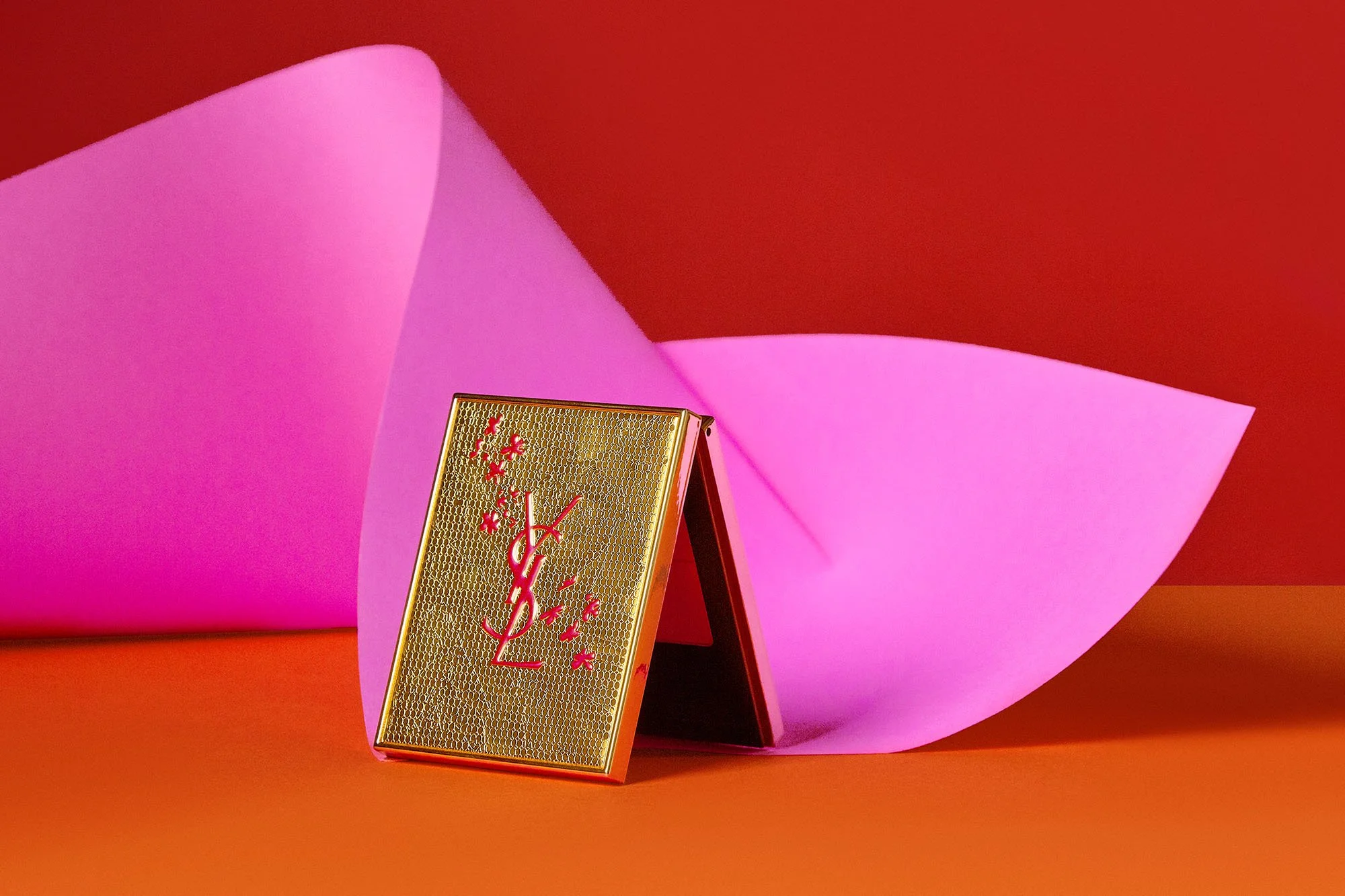 A gold and pink Yves Saint Laurent makeup compact placed on an orange surface, with pink paper resembling lips in the background.