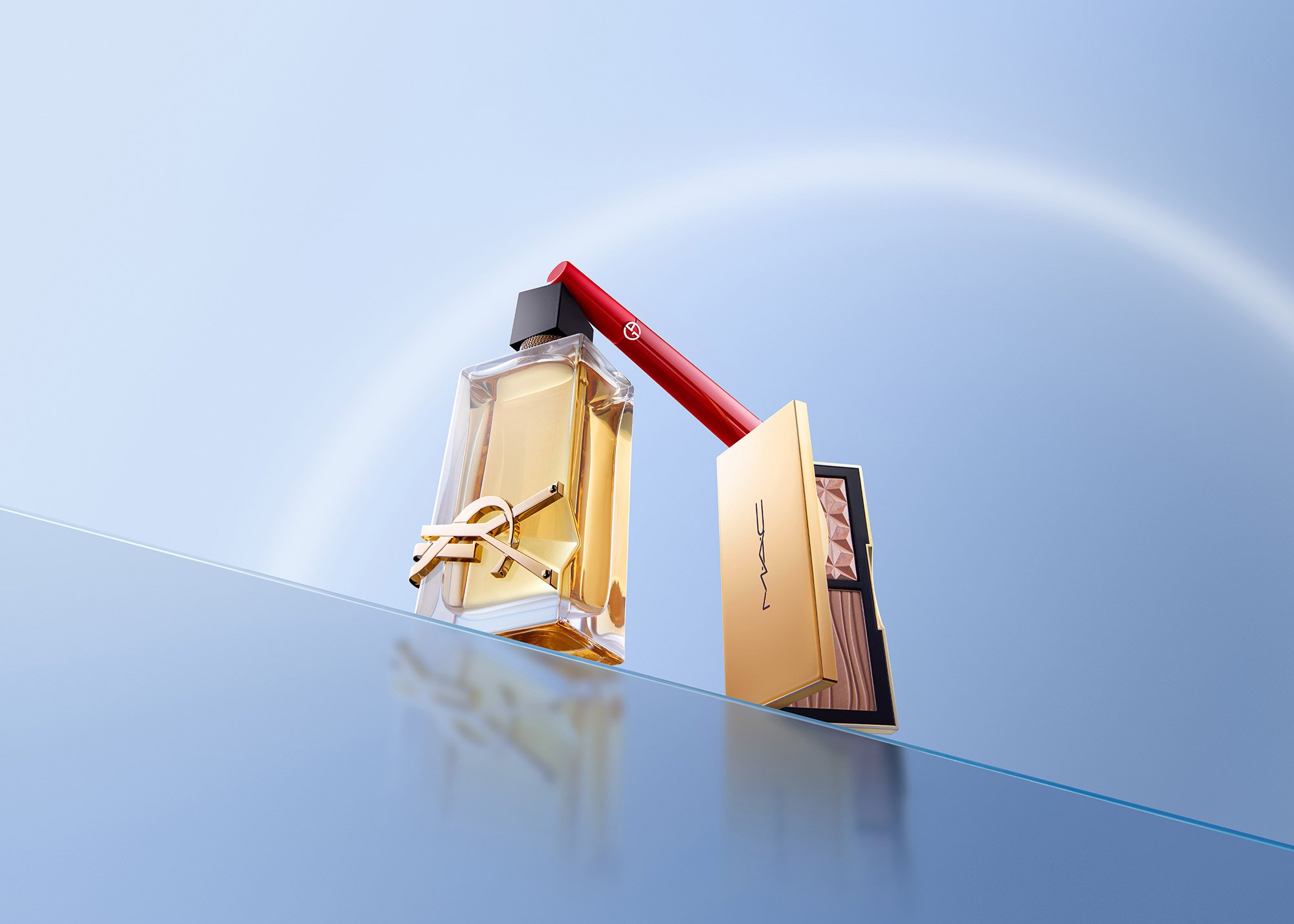 A perfume bottle, a MAC makeup palette, and a MAC lipstick with the cap off and the lipstick extended, arranged on a flat surface against a clear blue sky background.