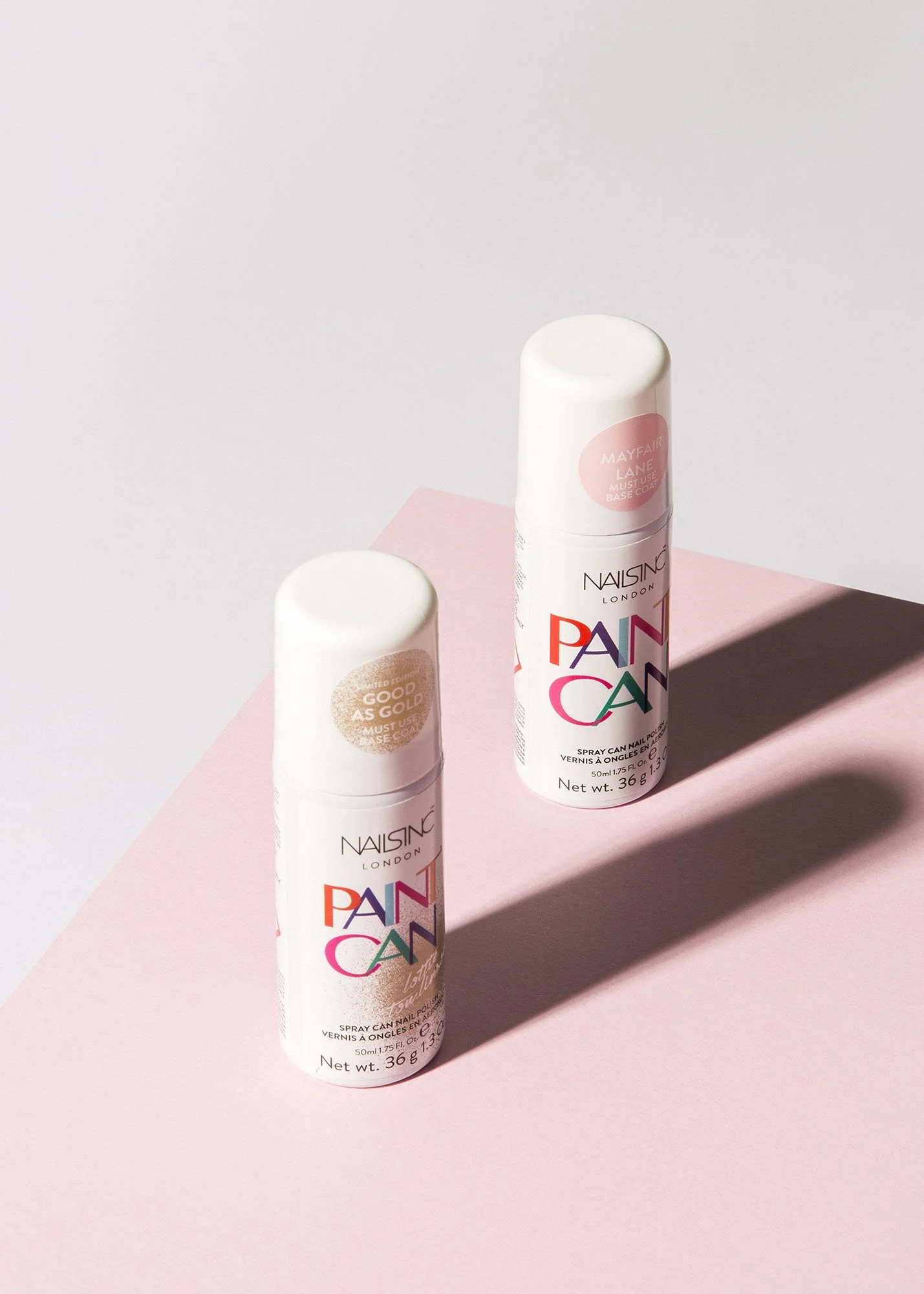 Two bottles of nail spray paint on a pink surface with soft shadows and a plain light background.