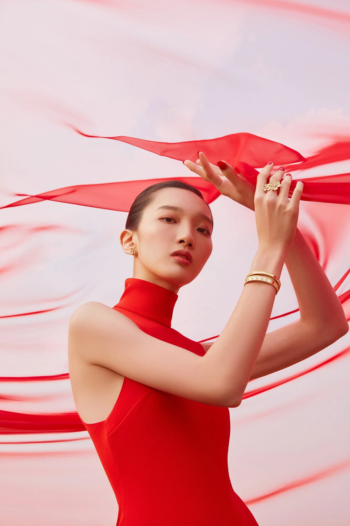 A woman wearing a red sleeveless turtleneck dress and gold jewelry, holding a flowing red scarf against a soft pink and white background.