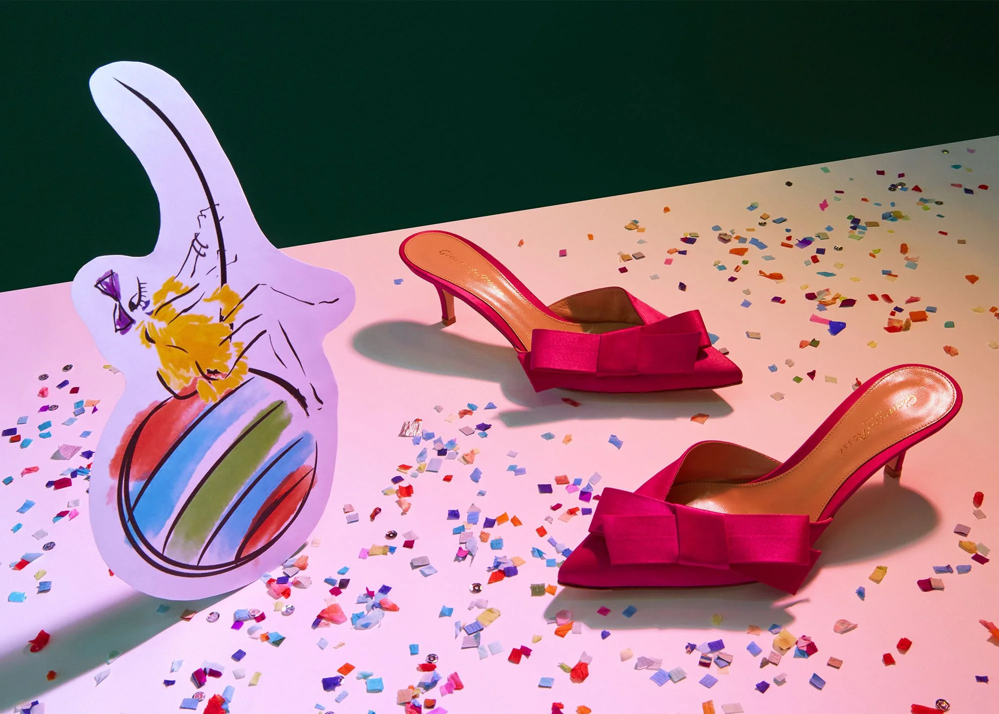Pink high-heeled mules with bows on pink and green background, colorful confetti scatter, paper cutout of a colorful balloon with a yellow cat wearing a purple bow.