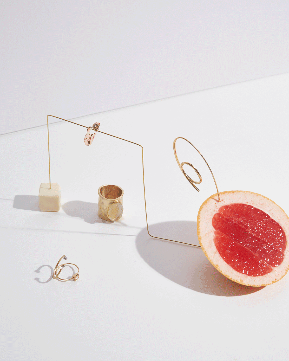 Arrangement of jewelry pieces including rings and earrings, a gold ring, and earrings, displayed on minimalist structures with a grapefruit half on the side, all set on a white background with shadows.