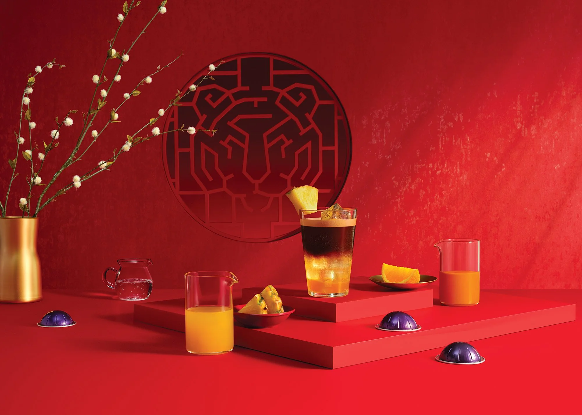 Red background with a golden vase holding white flowering branches on the left, a coffee-like drink topped with pineapple in the center, and glasses of orange juice and slices of orange on rectangular platforms. Small purple objects are scattered on 
