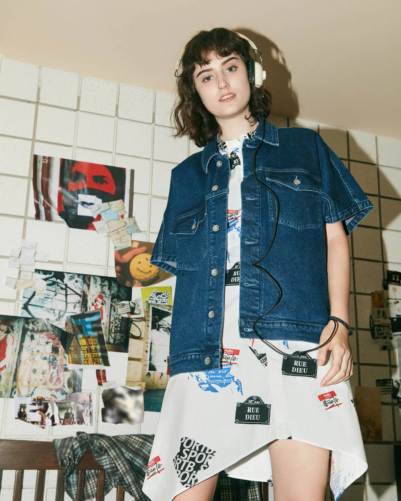 A young woman with curly brown hair wearing a denim jacket over a printed dress and headphones, standing in front of a wall decorated with posters and pictures.
